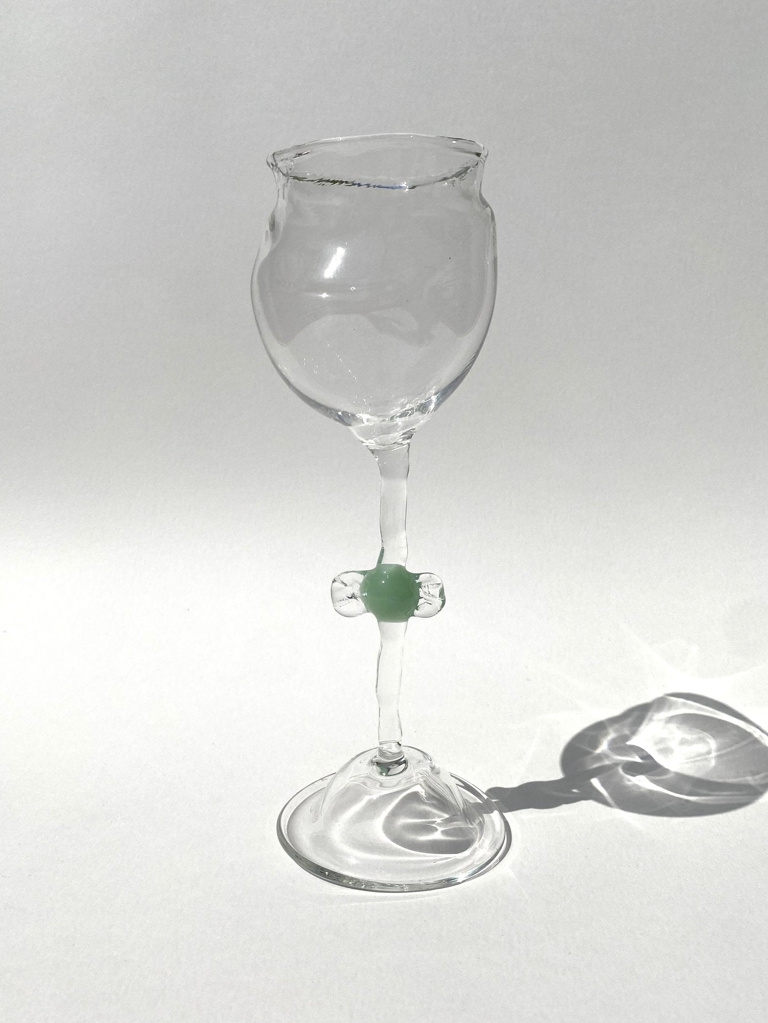A clear mouth-blown wine glasses by Vivian Alarcón with a green candy-shaped embellishment on the stem, displayed against a light background.