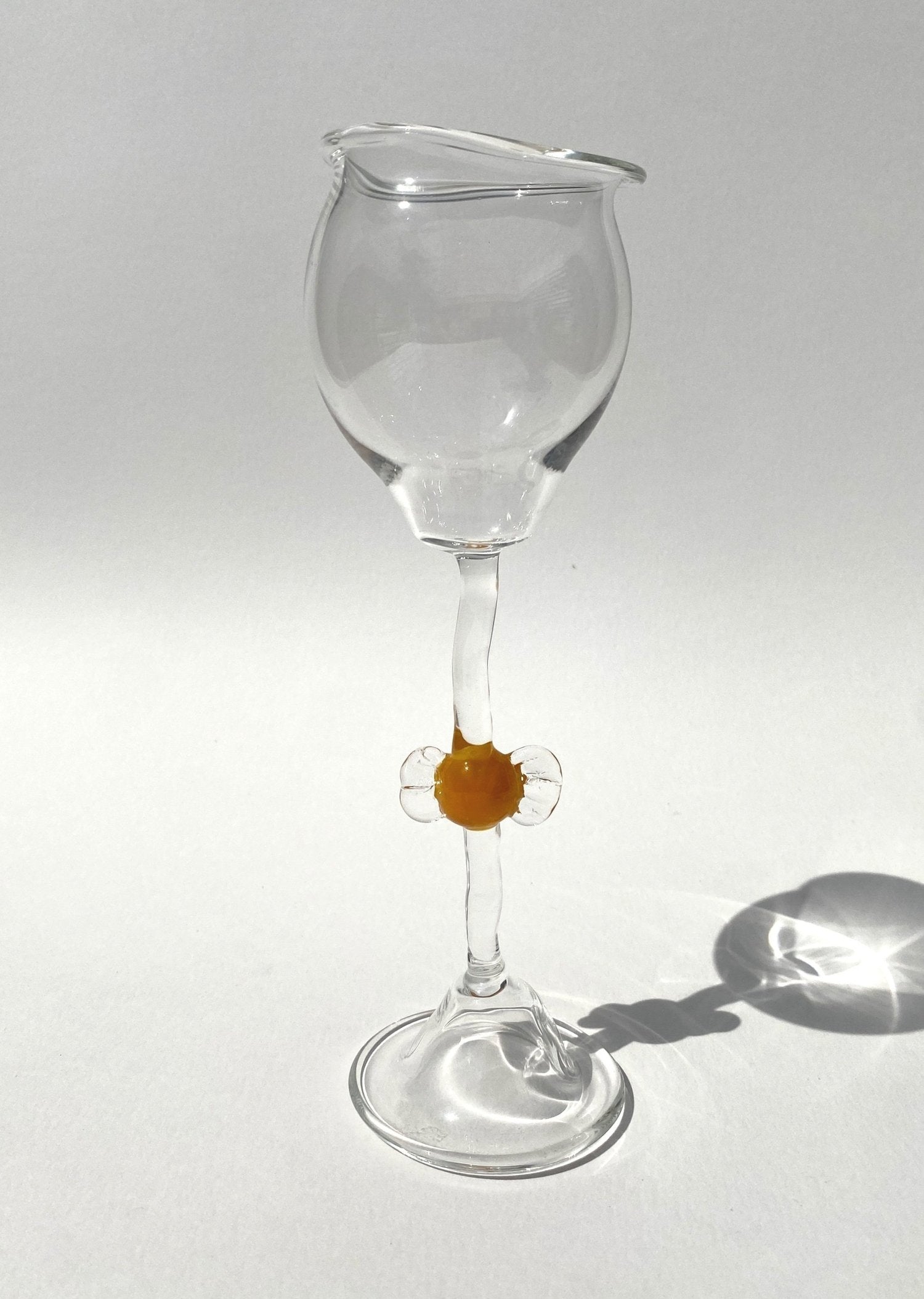 A clear mouth-blown wine glasses by Vivian Alarcón with an orange candy-shaped embellishment on the stem, displayed against a light background.