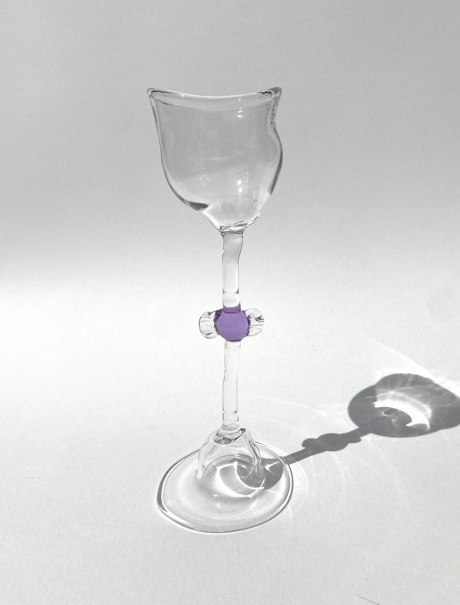 A clear mouth-blown wine glasses by Vivian Alarcón with a purple candy-shaped embellishment on the stem, displayed against a light background.