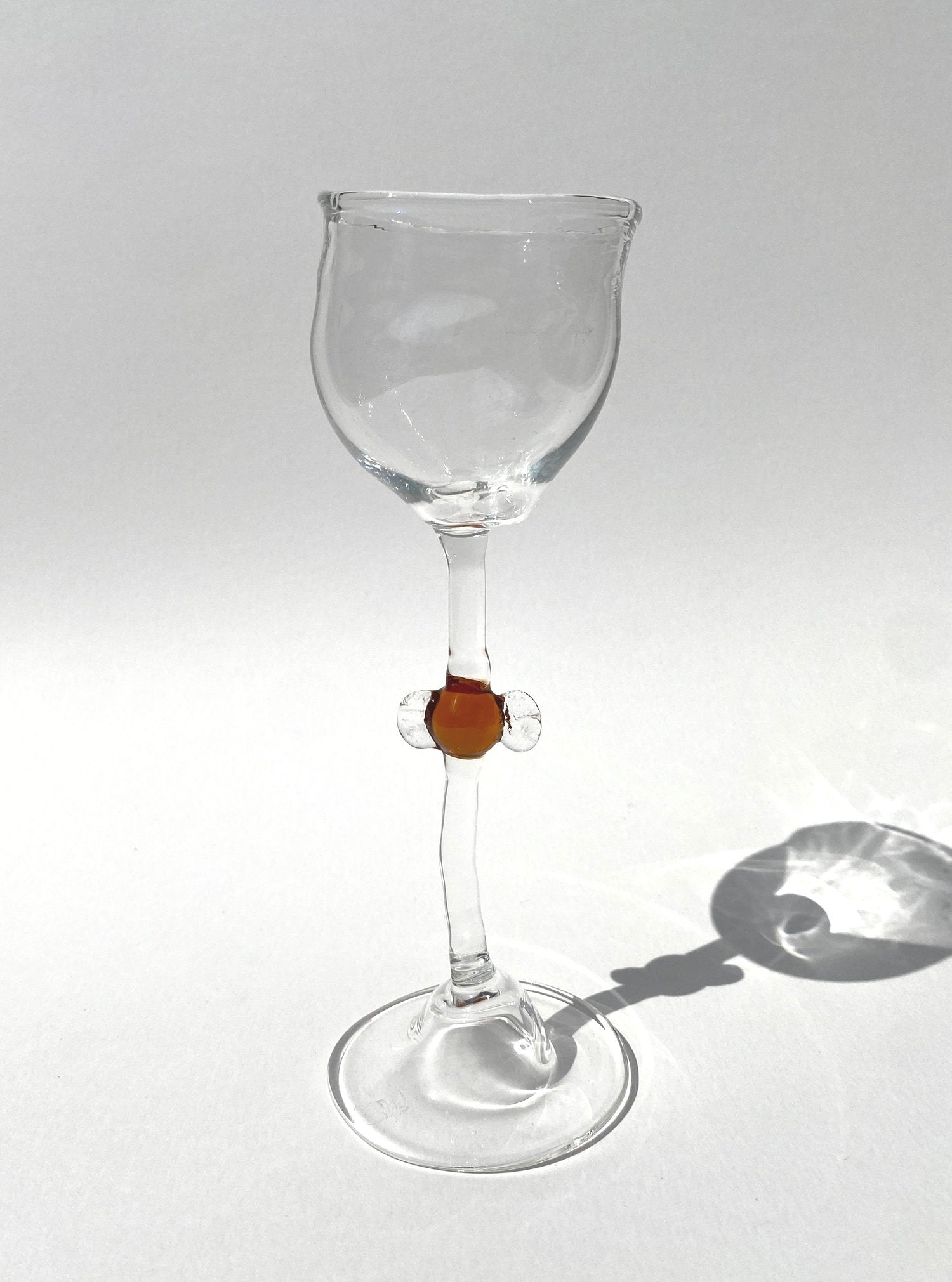 A clear mouth-blown wine glasses by Vivian Alarcón with a brown candy-shaped embellishment on the stem, displayed against a light background.