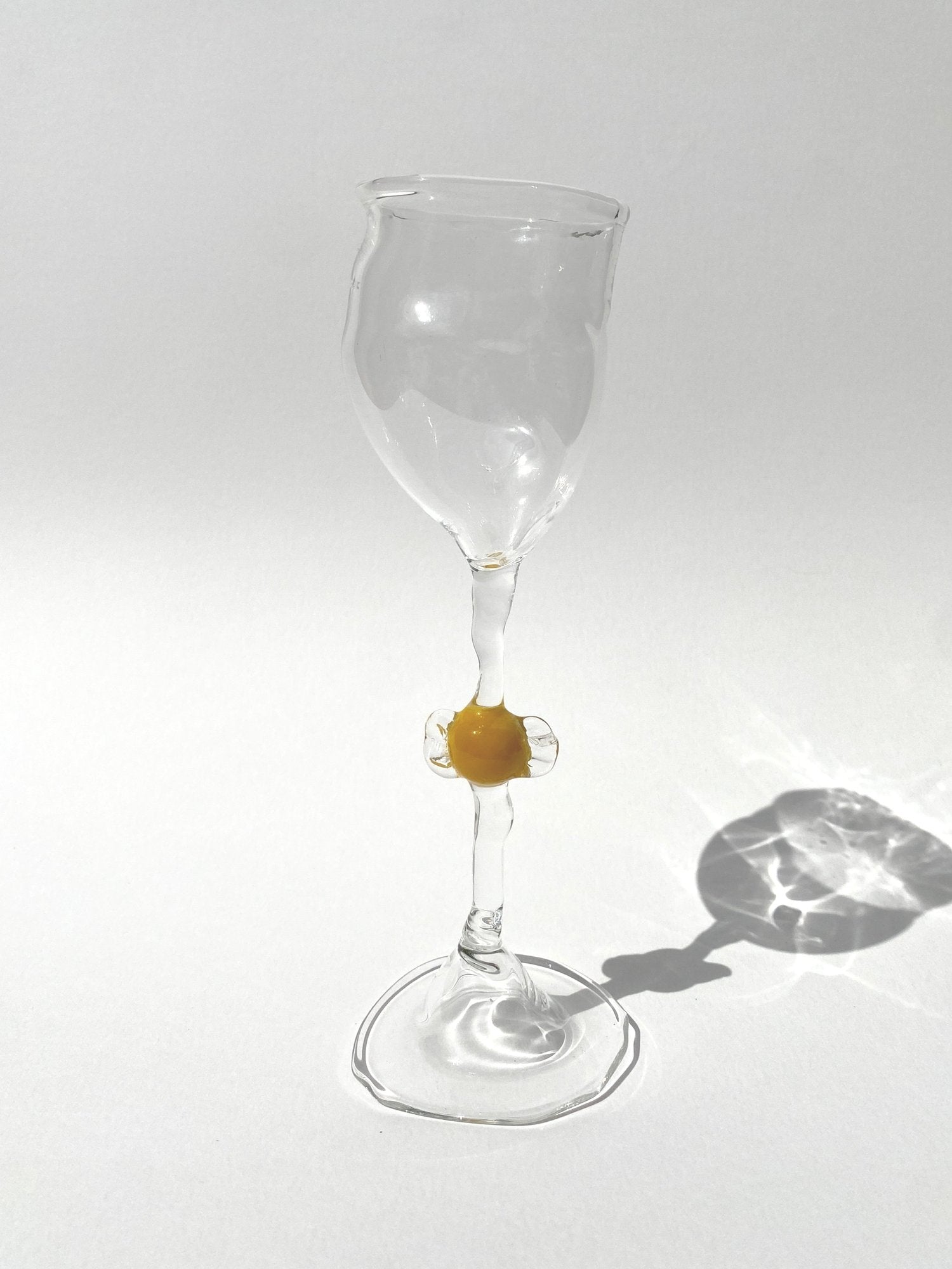 A clear mouth-blown wine glasses by Vivian Alarcón with a brown candy-shaped embellishment on the stem, displayed against a light background.