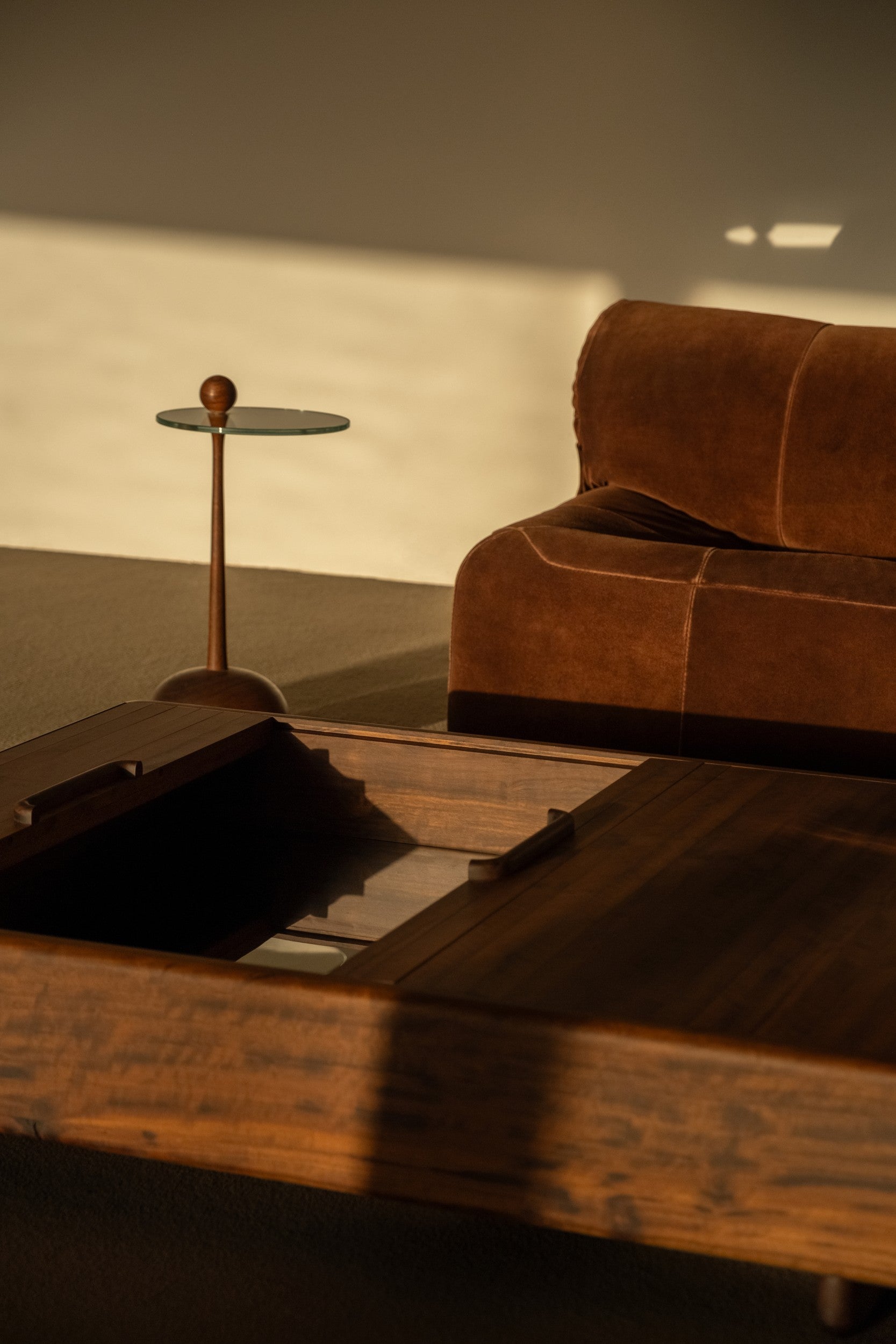 Wooden side table with glass top by Estudio Prosa in a warm living room setting with a brown, suede sofa and a wooden coffee table.