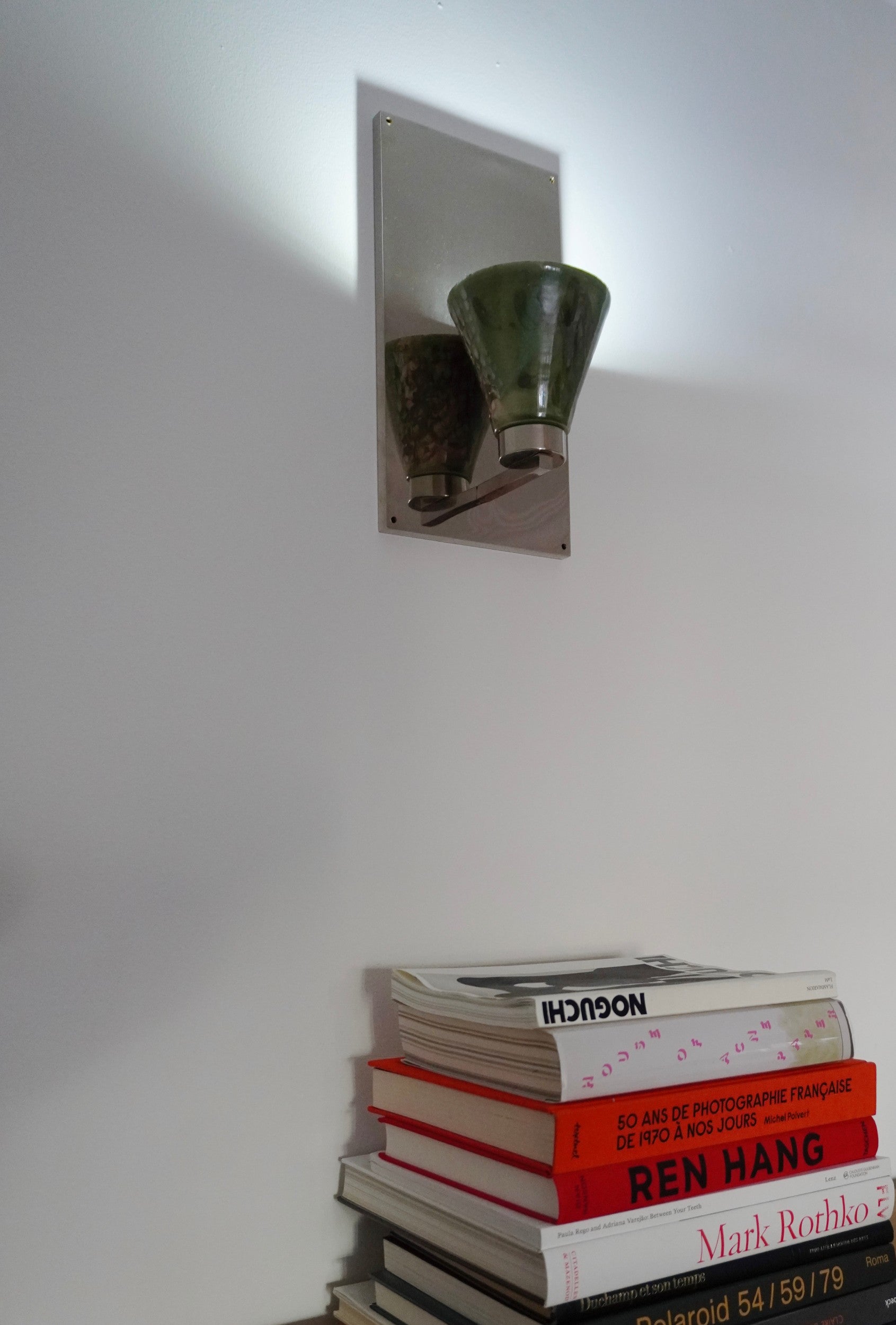 Vetra Lamp by Marta Cypel, mounted on a white wall above a stack of books.