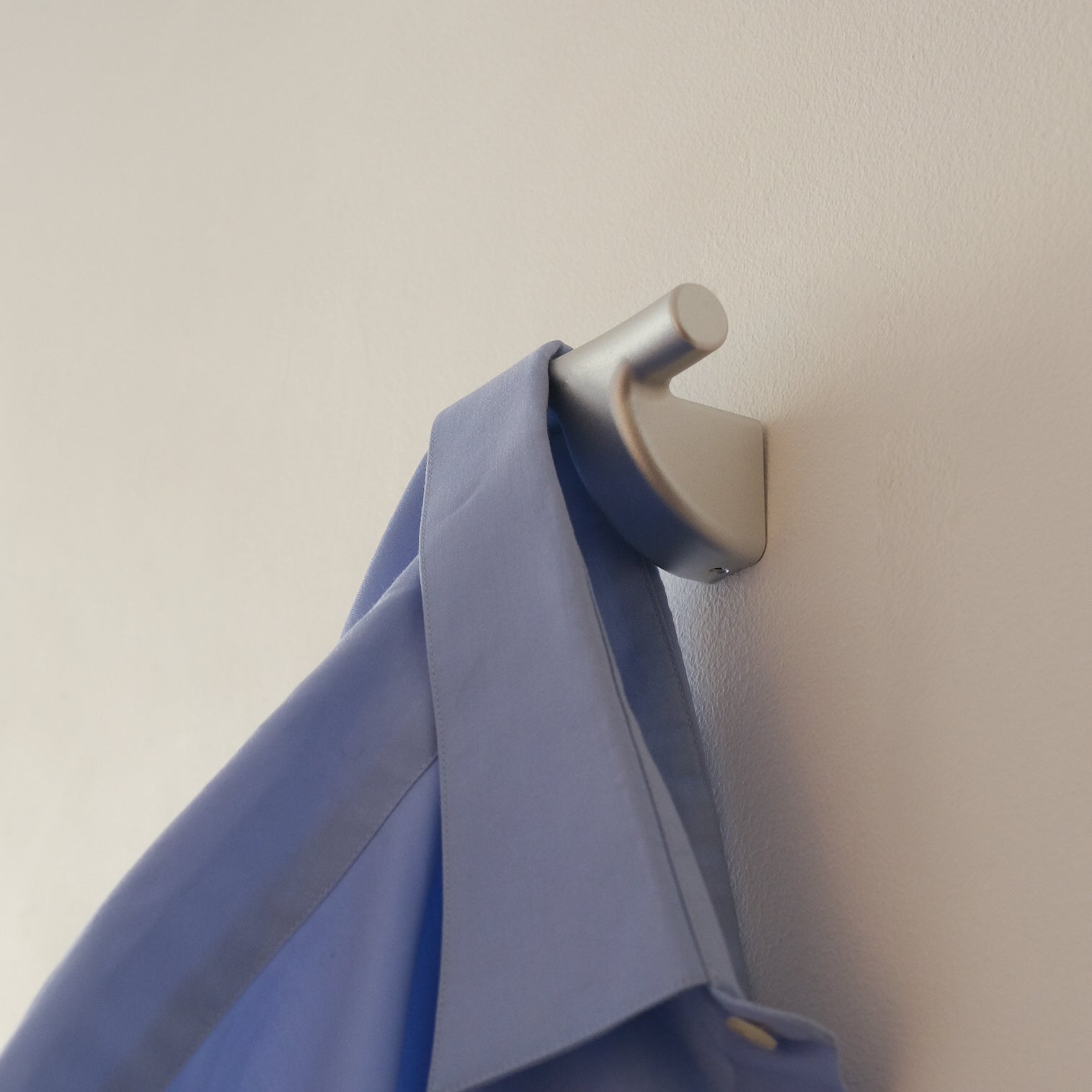 Silver mooring hook by PARCTA mounted on a neutral wall with a blue shirt hooked to it.