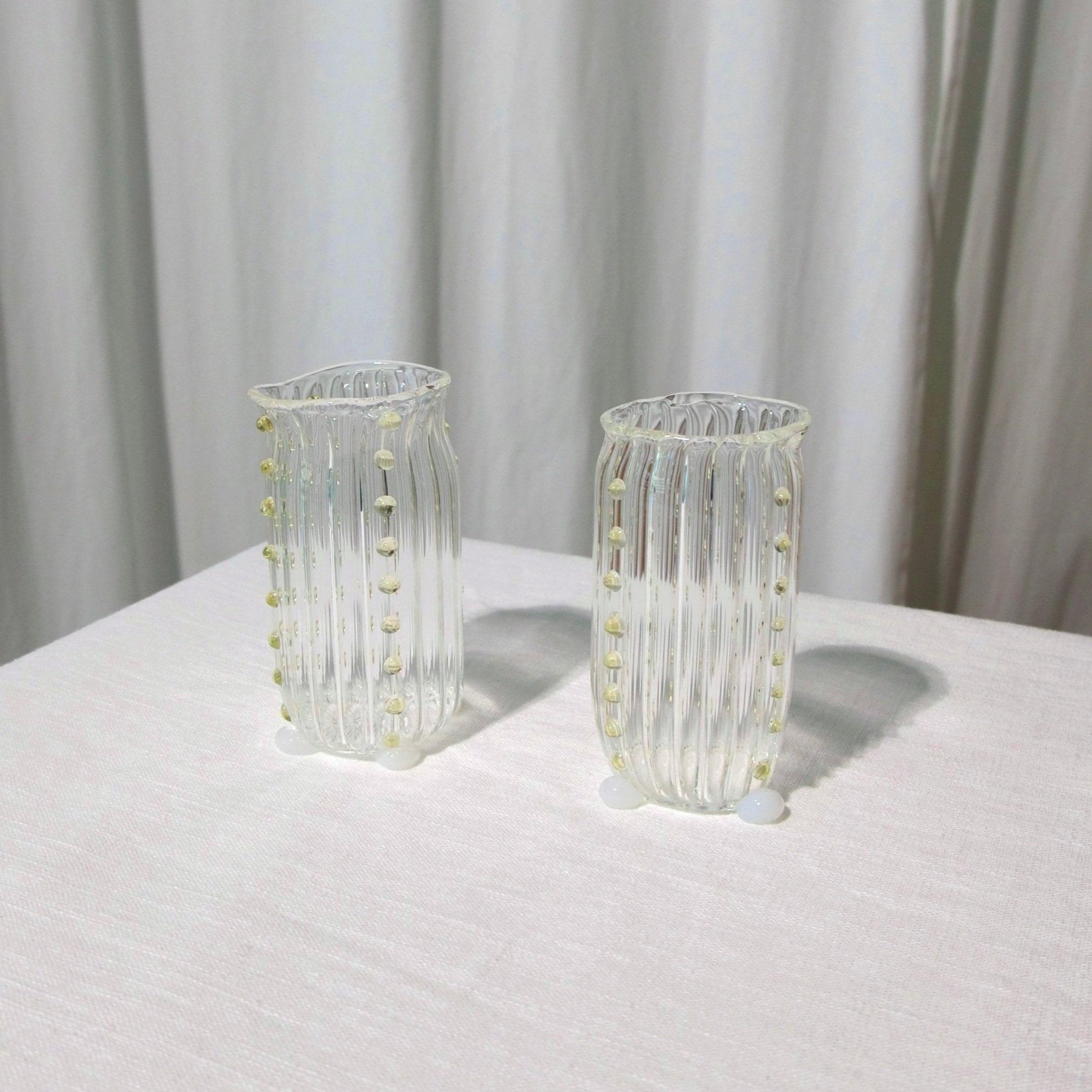Two mouth-blown, decorative water glasses by Vivian Alarcón on a table with white table linen and neutral curtains in the background.