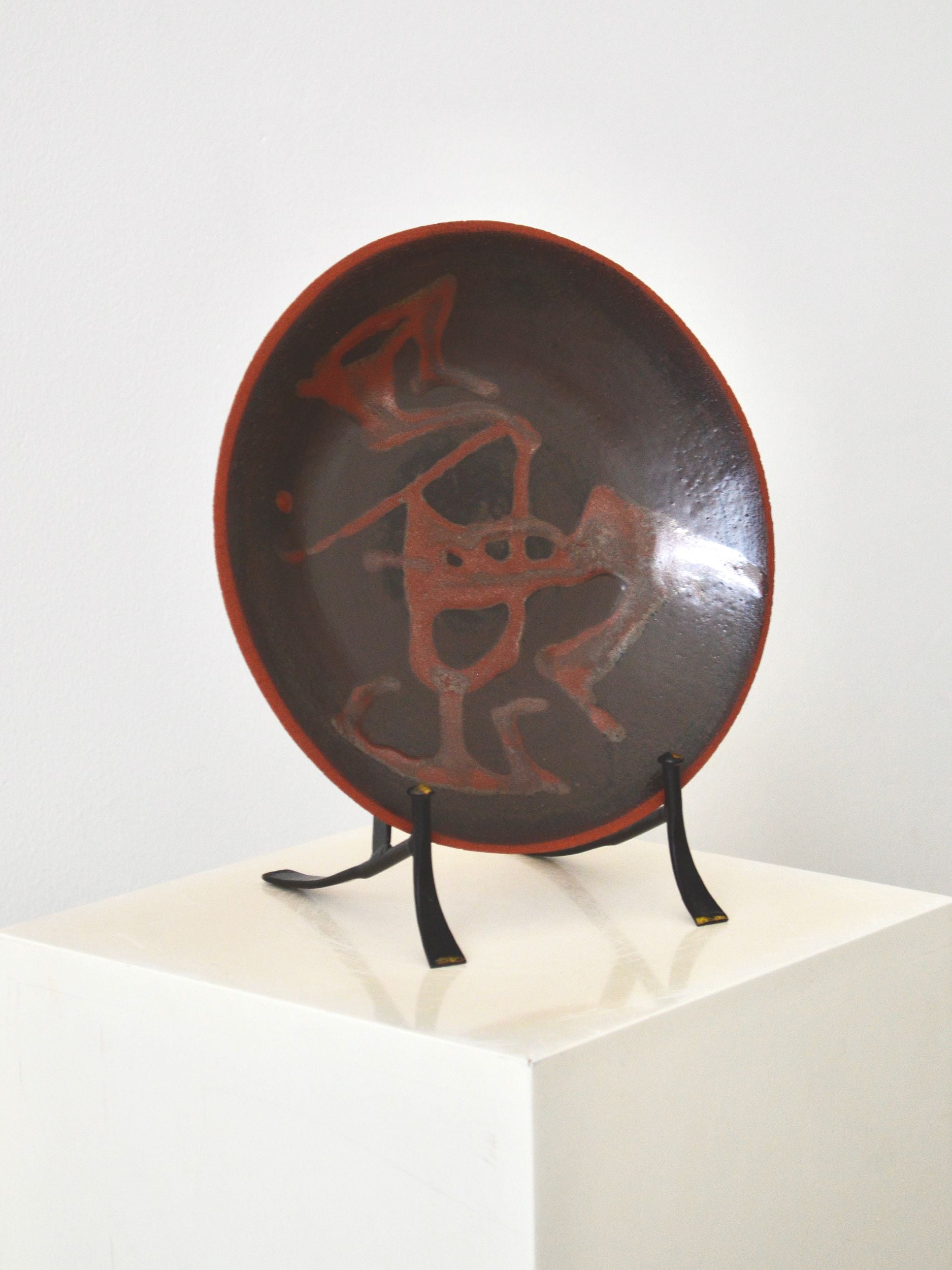 A decorative ceramic plate with an organic swirling pattern in shades of brown by Zeynep Boyan. The plate is positioned on a stand on a white plinth with a white background.