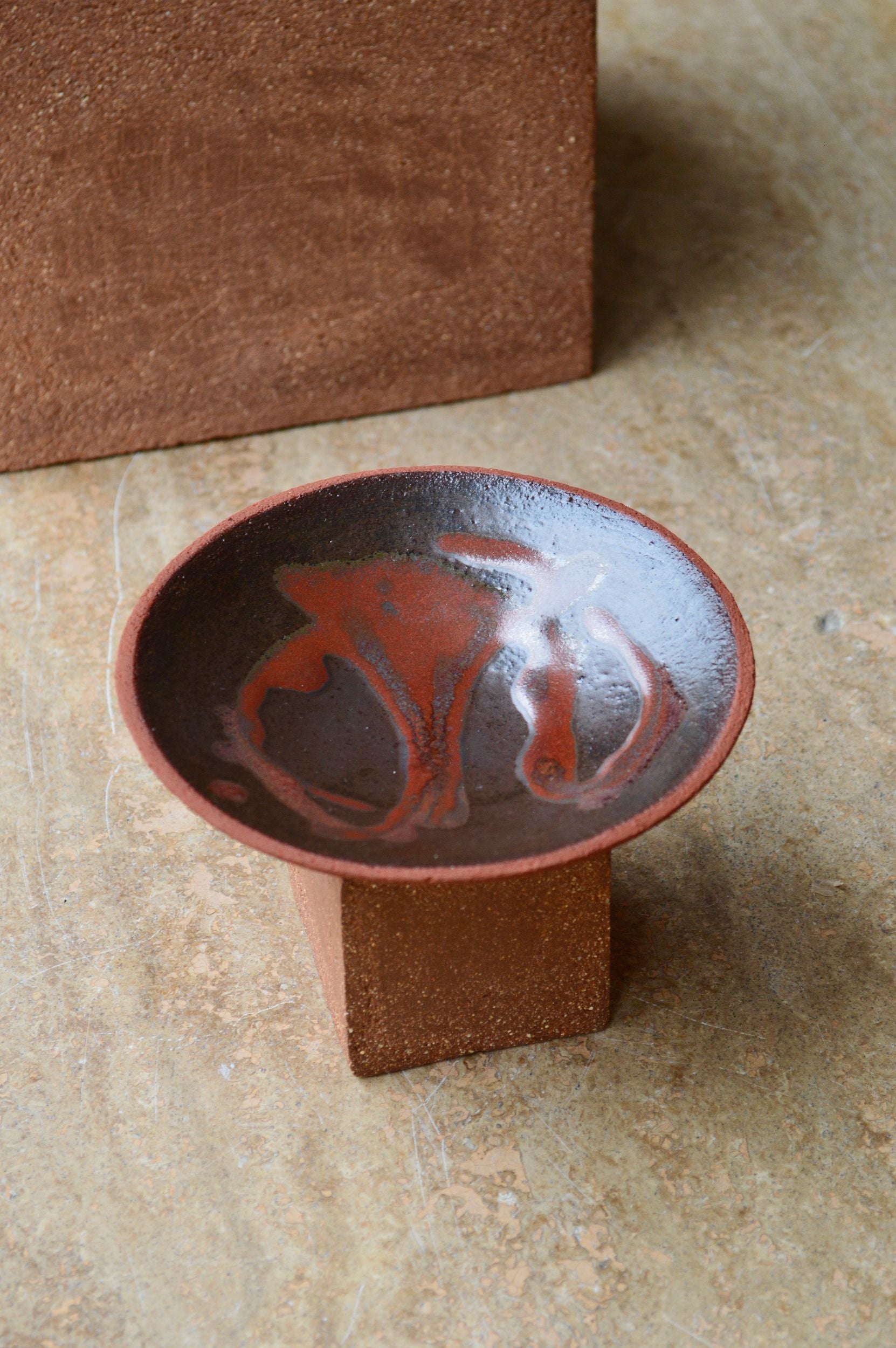 A decorative ceramic plate with an organic swirling pattern in shades of brown by Zeynep Boyan. The plate is positioned on a brick on neutral flooring.