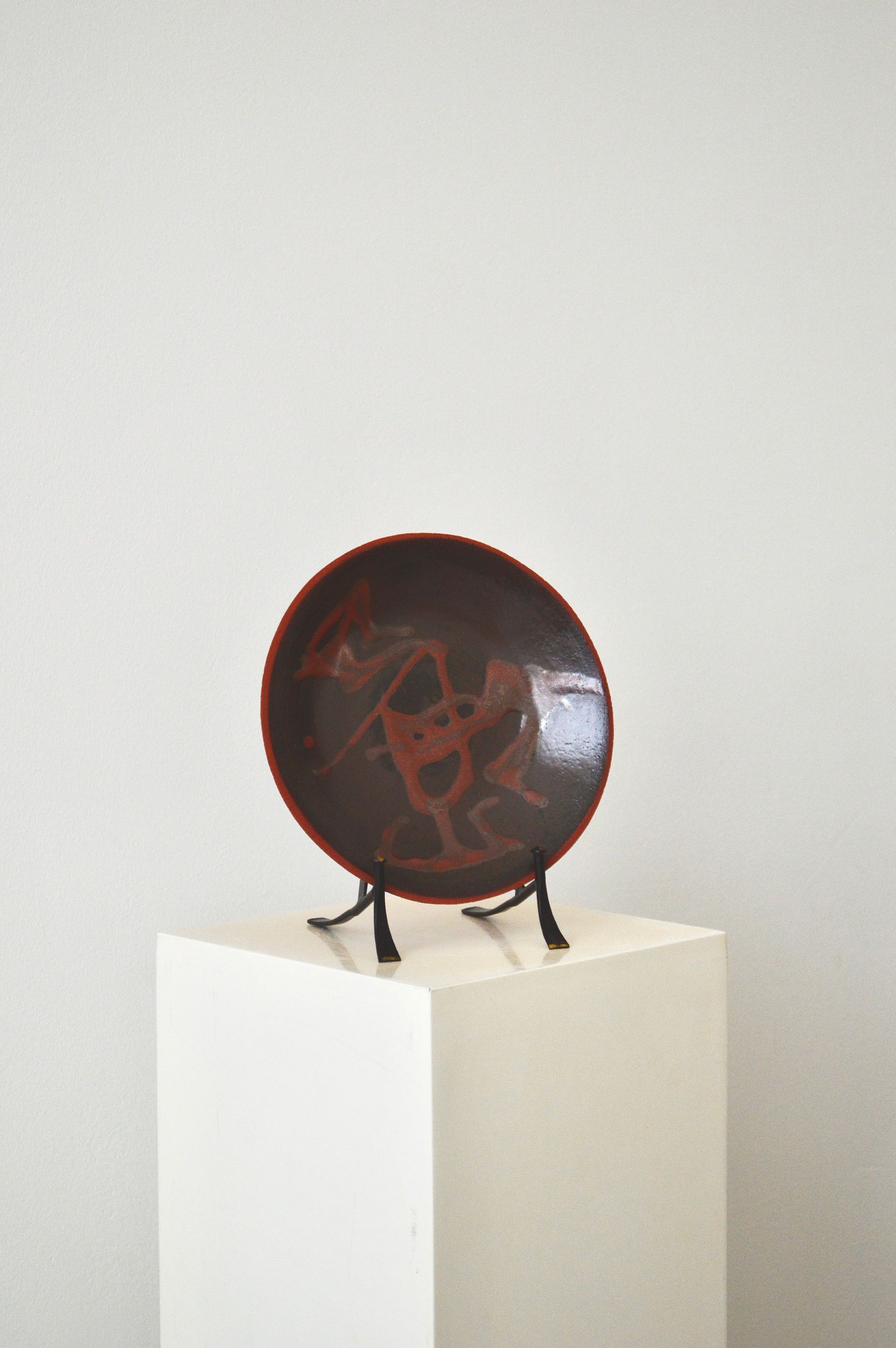 A decorative ceramic plate with an organic swirling pattern in shades of brown by Zeynep Boyan. The plate is positioned on a stand on a white plinth with a white background.
