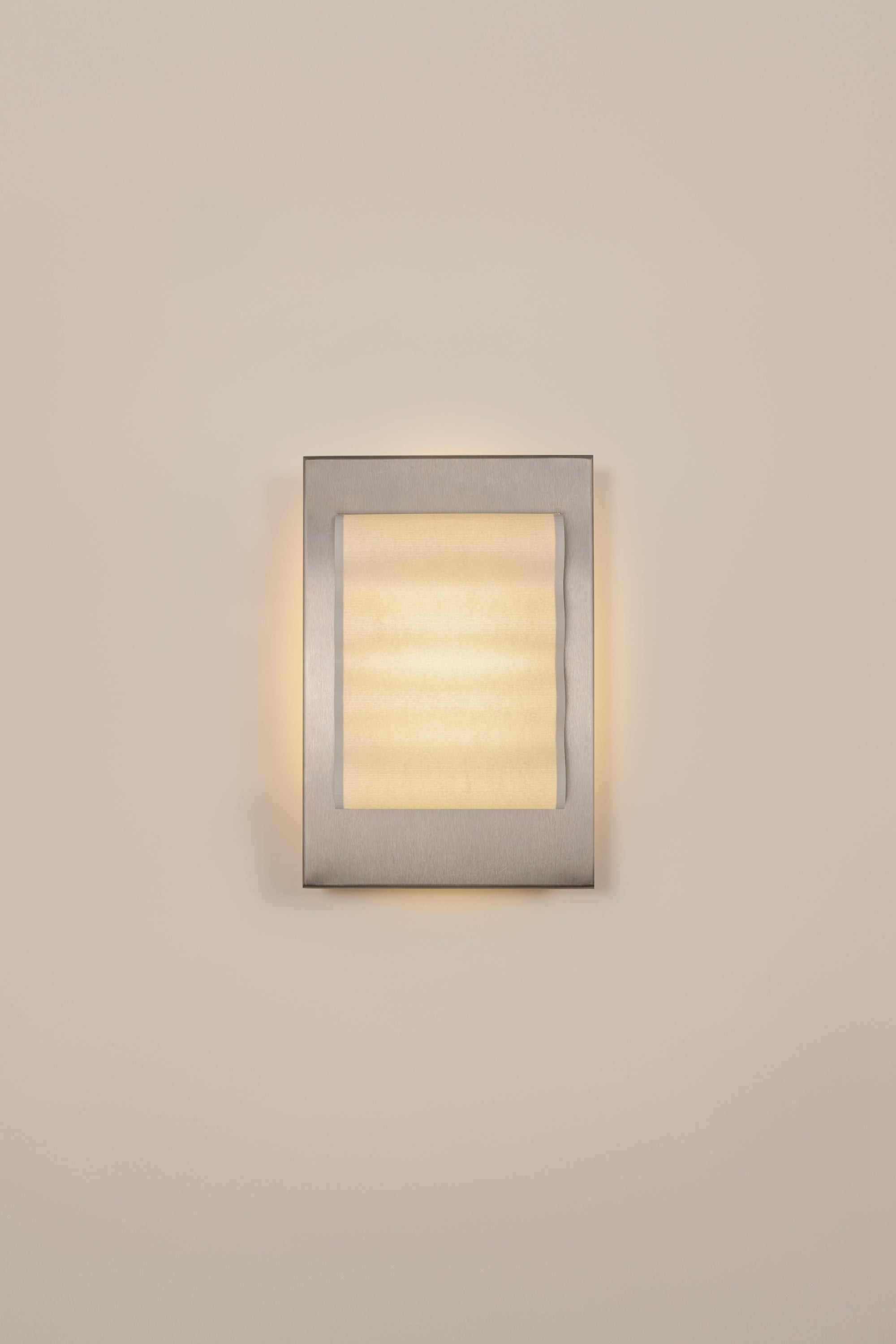 Framed wall light fixture in raw stainless steel with a paper shade by Violaine d'Harcourt on a beige wall with light on.