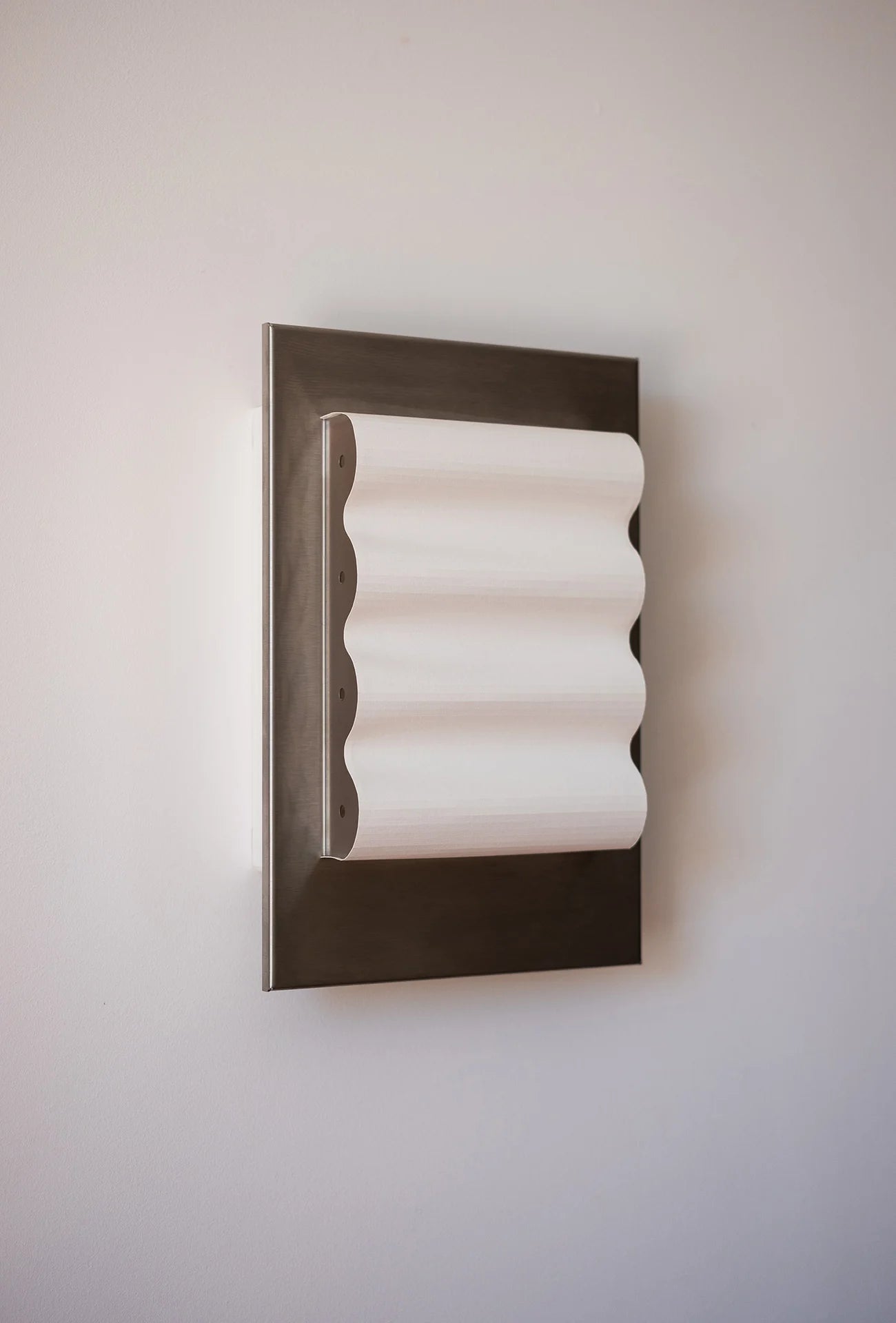 Framed wall light fixture in raw stainless steel with a paper shade by Violaine d'Harcourt on a beige wall with light off.