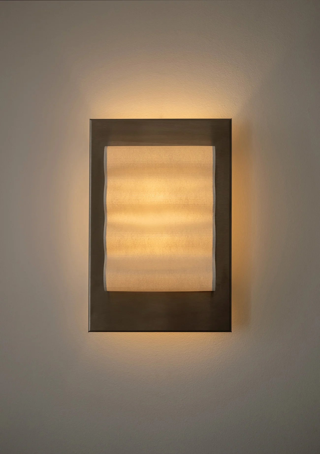 Framed wall light fixture in raw stainless steel with a paper shade by Violaine d'Harcourt on a beige wall with light on.