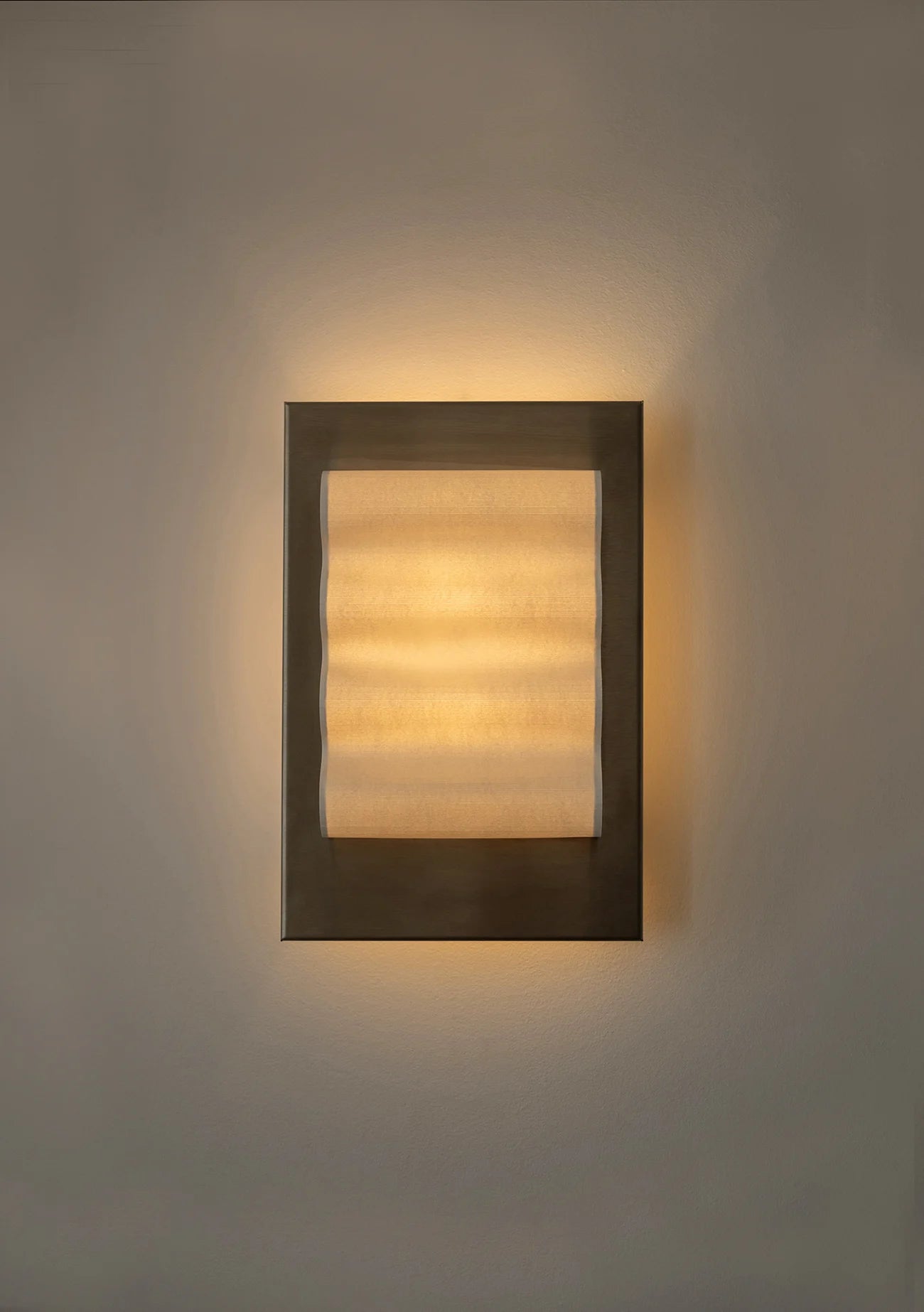 Framed wall light fixture in raw stainless steel with a paper shade by Violaine d'Harcourt on a beige wall with light on.