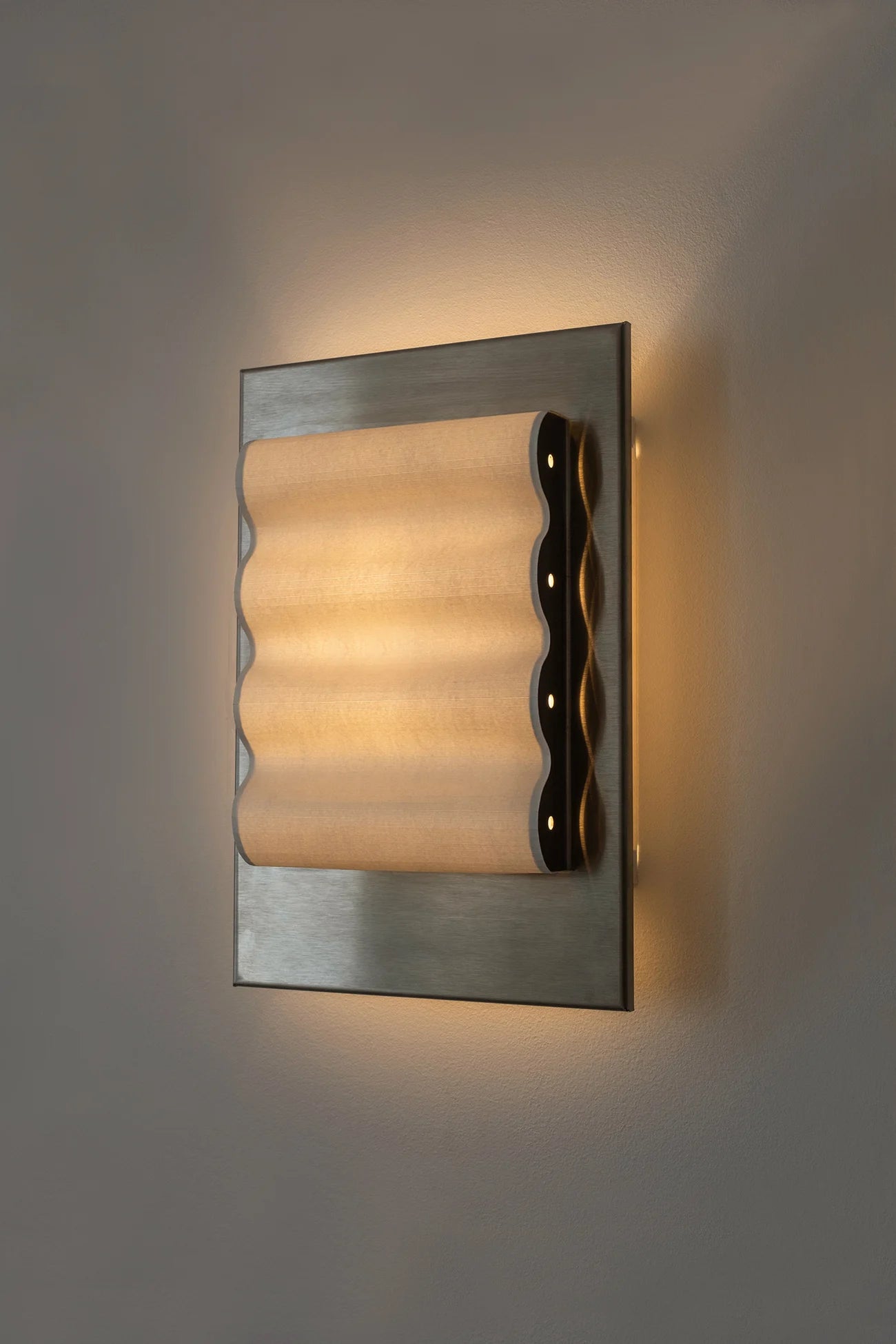 Framed wall light fixture in raw stainless steel with a paper shade by Violaine d'Harcourt on a beige wall with light on.