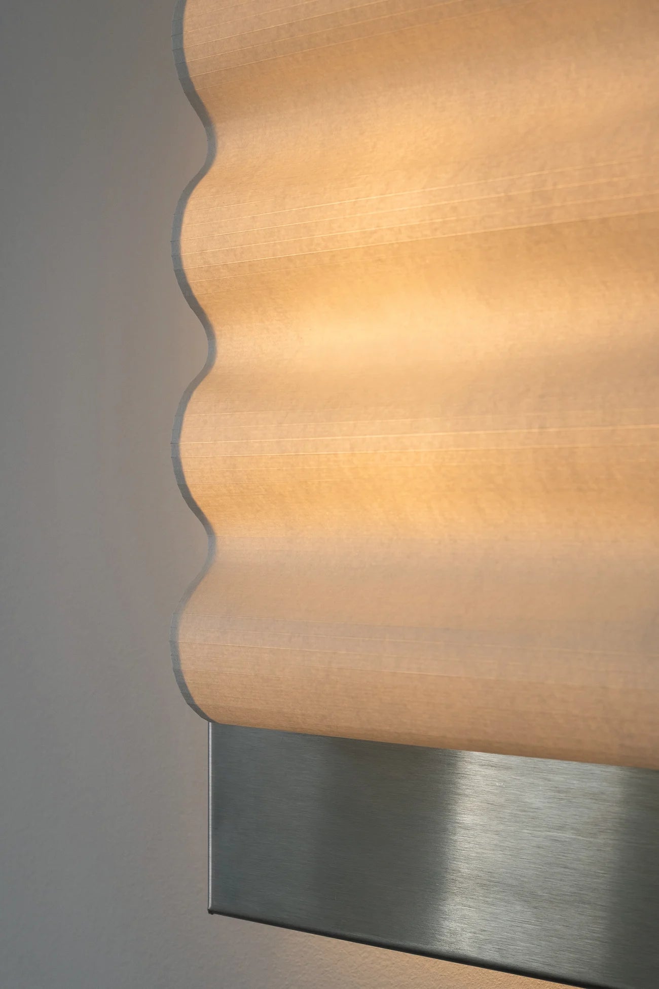 Close up of framed wall light fixture in raw stainless steel with a paper shade by Violaine d'Harcourt on a beige wall with light on.