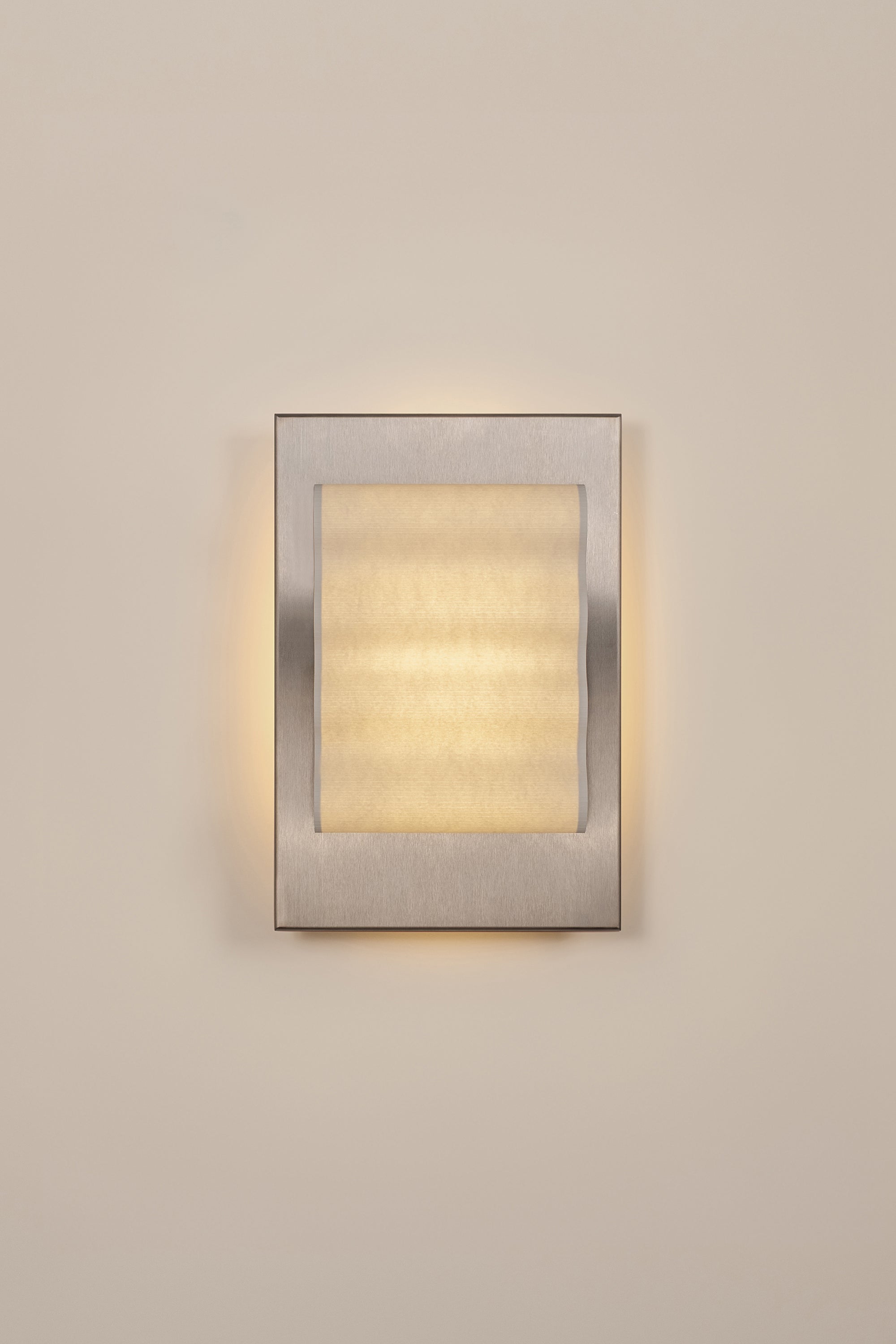 Framed wall light fixture in raw stainless steel with a paper shade by Violaine d'Harcourt on a beige wall with light on.