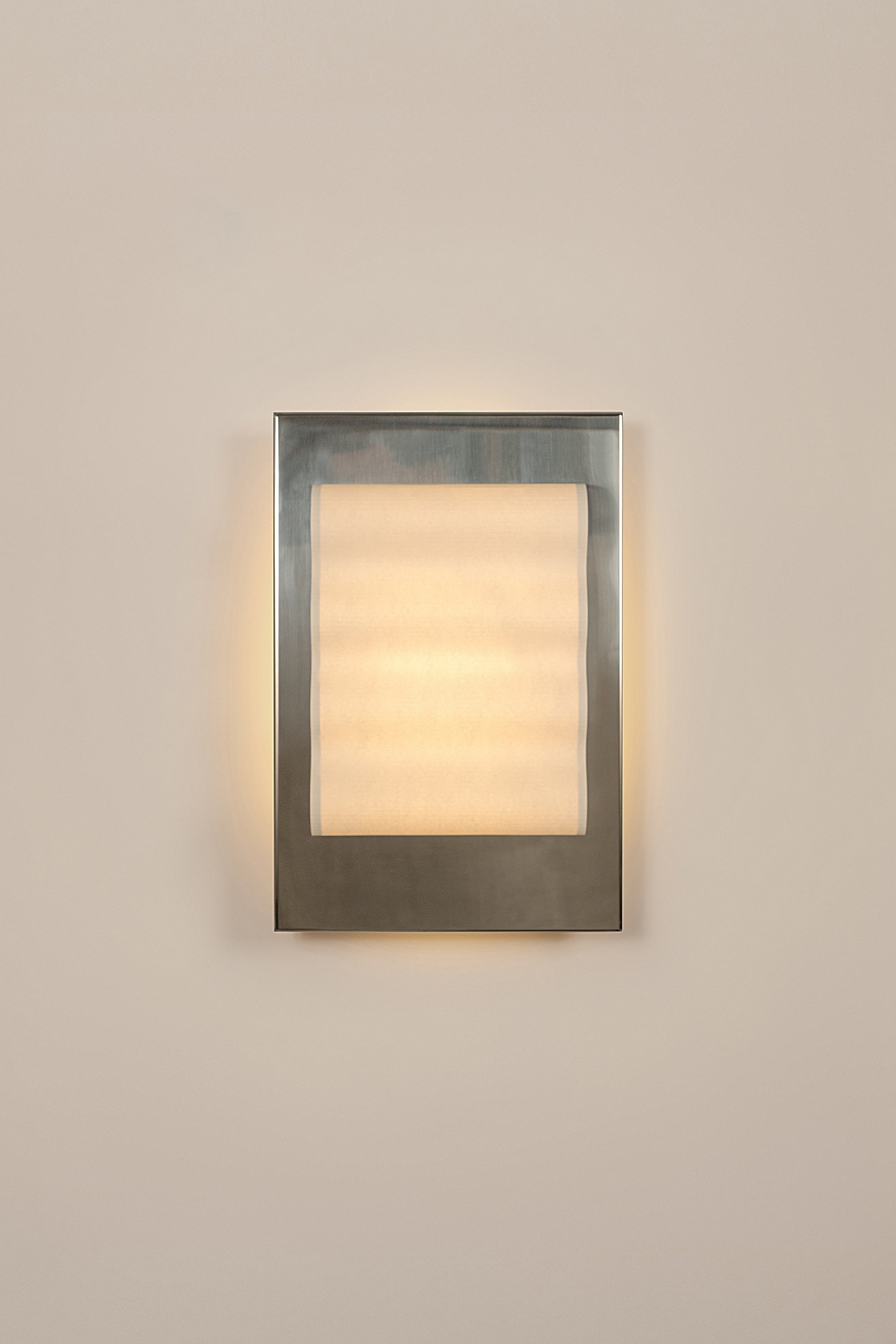 Framed wall light fixture in polished stainless steel with a paper shade by Violaine d'Harcourt on a beige wall with light on.