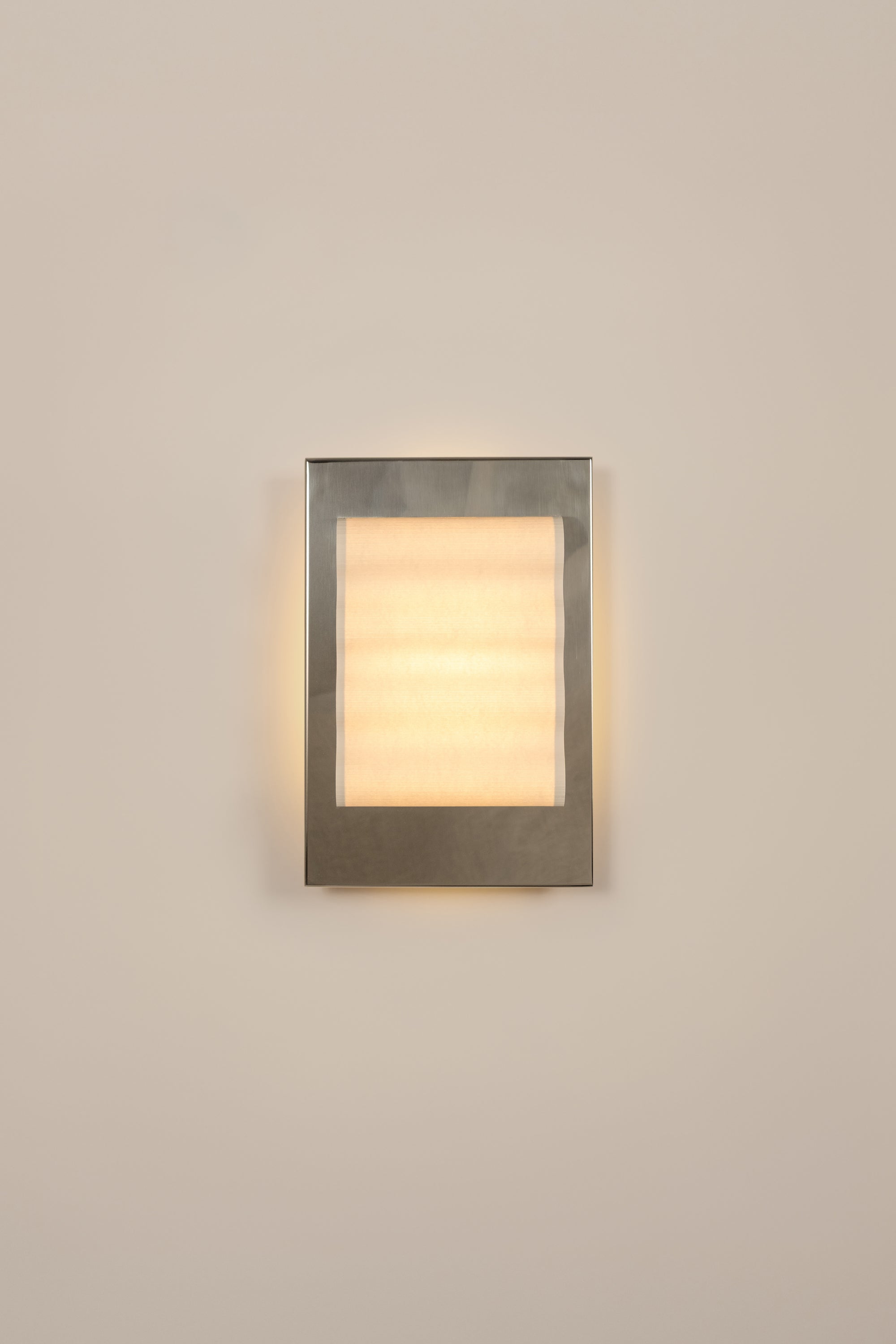 Framed wall light fixture in polished stainless steel with a paper shade by Violaine d'Harcourt on a beige wall with light on.