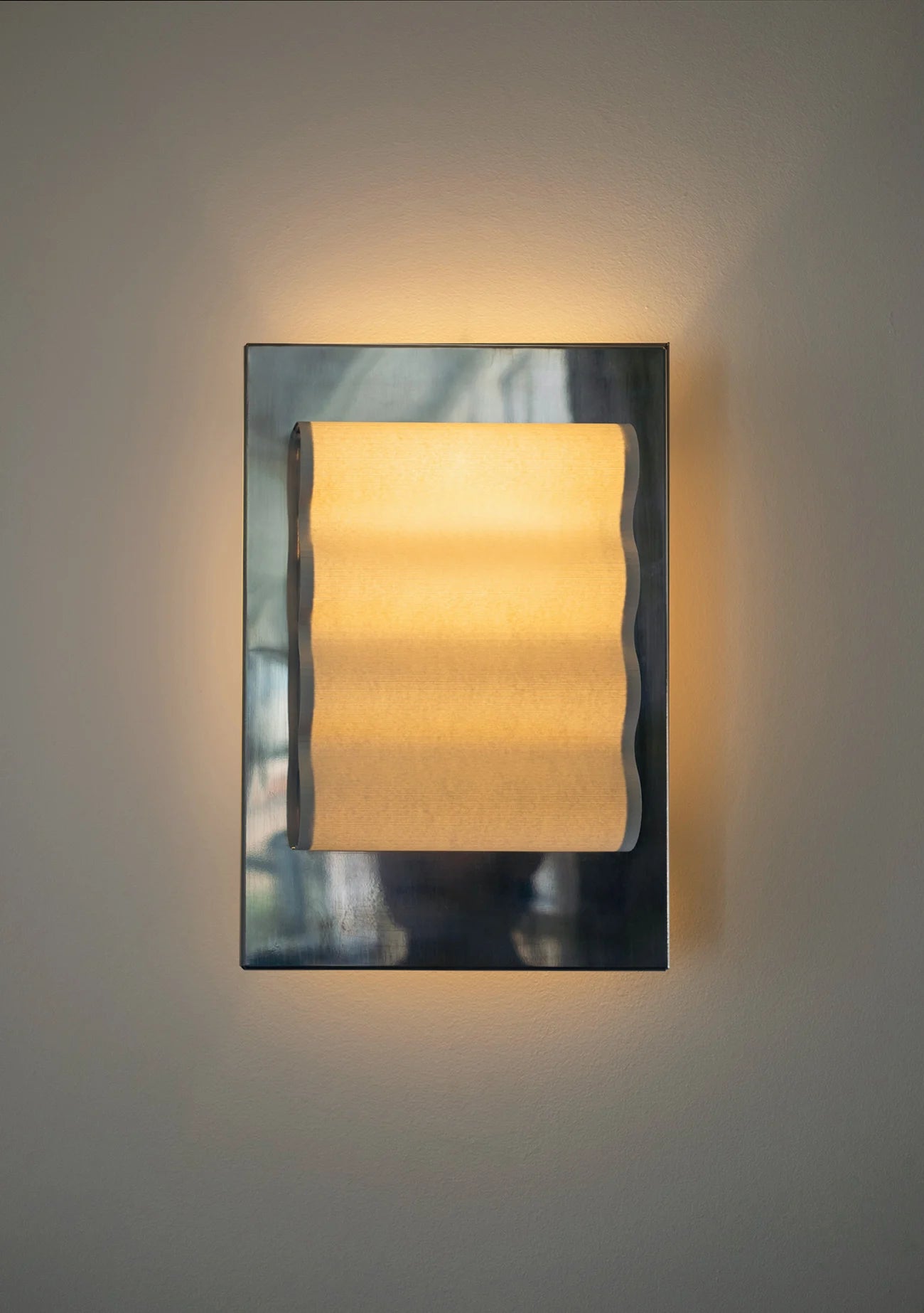 Framed wall light fixture in polished stainless steel with a paper shade by Violaine d'Harcourt on a beige wall with light on.