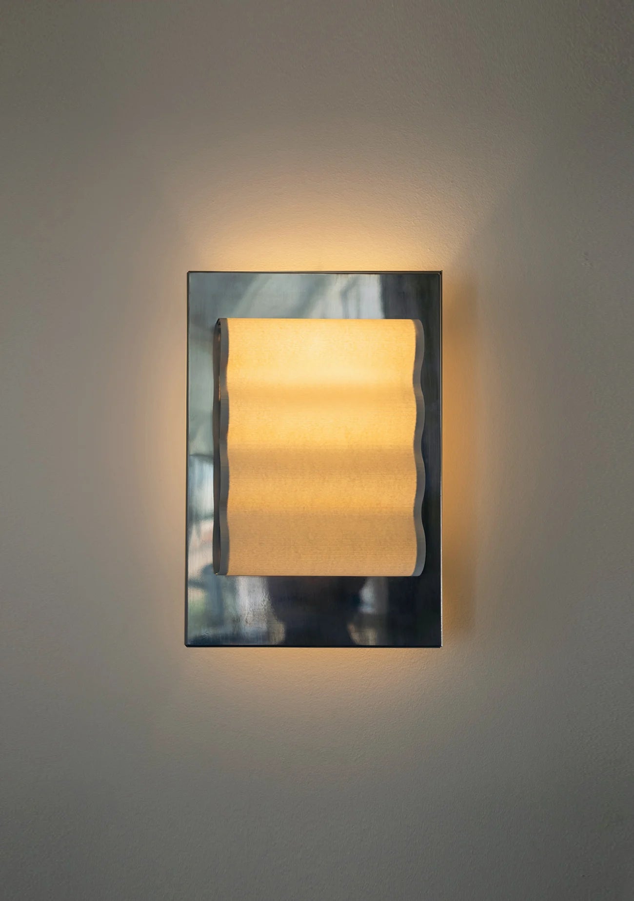Framed wall light fixture in polished stainless steel with a paper shade by Violaine d'Harcourt on a beige wall with light on.
