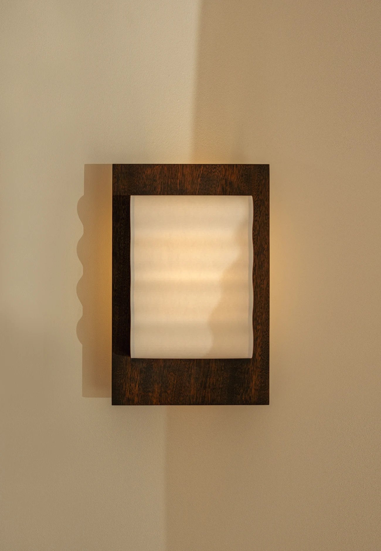 Framed wall light fixture in wood with a paper shade by Violaine d'Harcourt on a beige wall with light on.