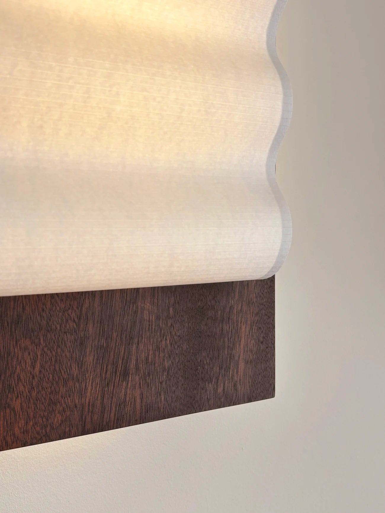 Close up of framed wall light fixture in wood with a paper shade by Violaine d'Harcourt on a beige wall with light on.