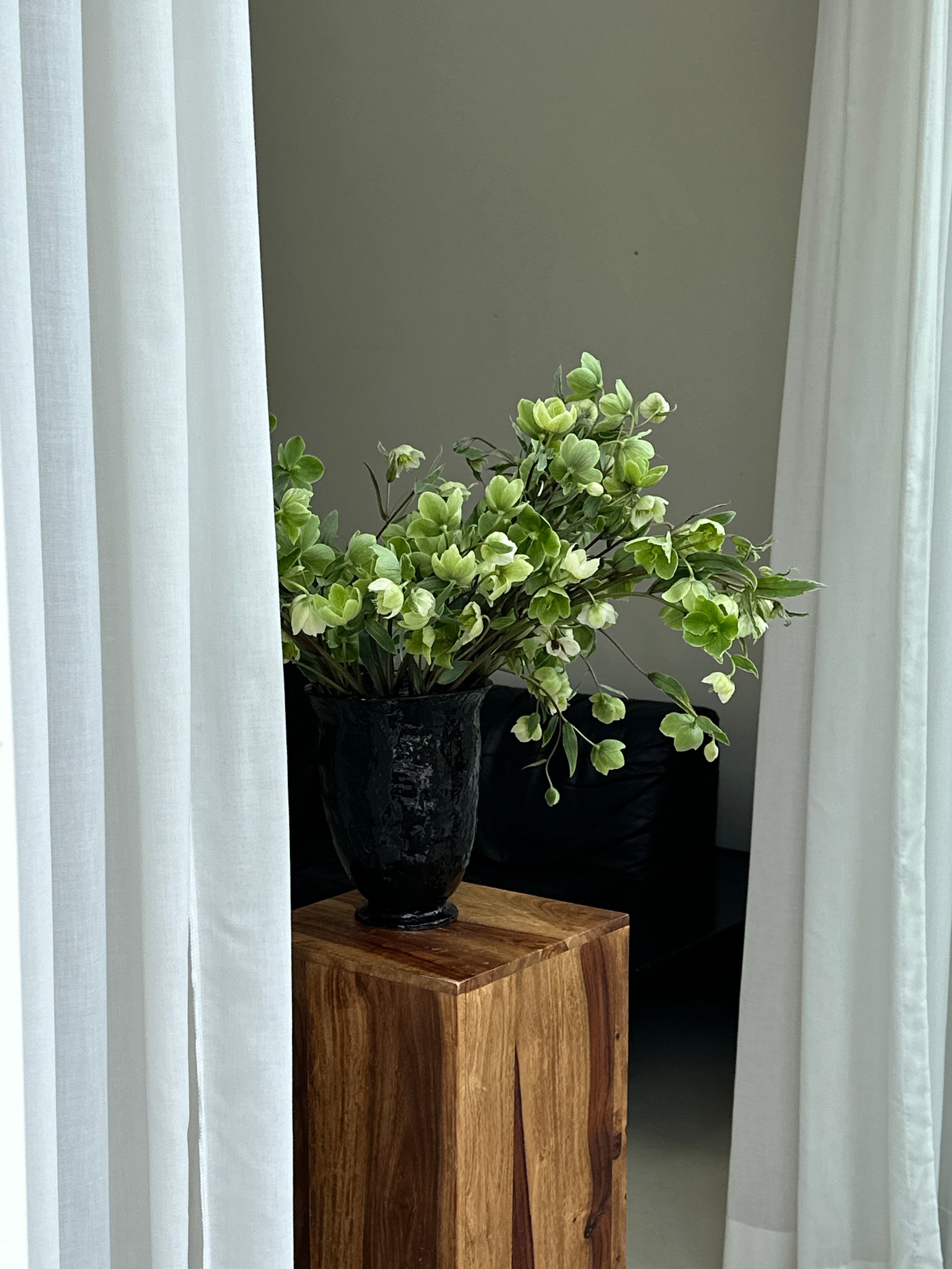 Black, glossy ceramic chalice vase by klangnapha on a wooden plinth. The vase is holding green flowers.