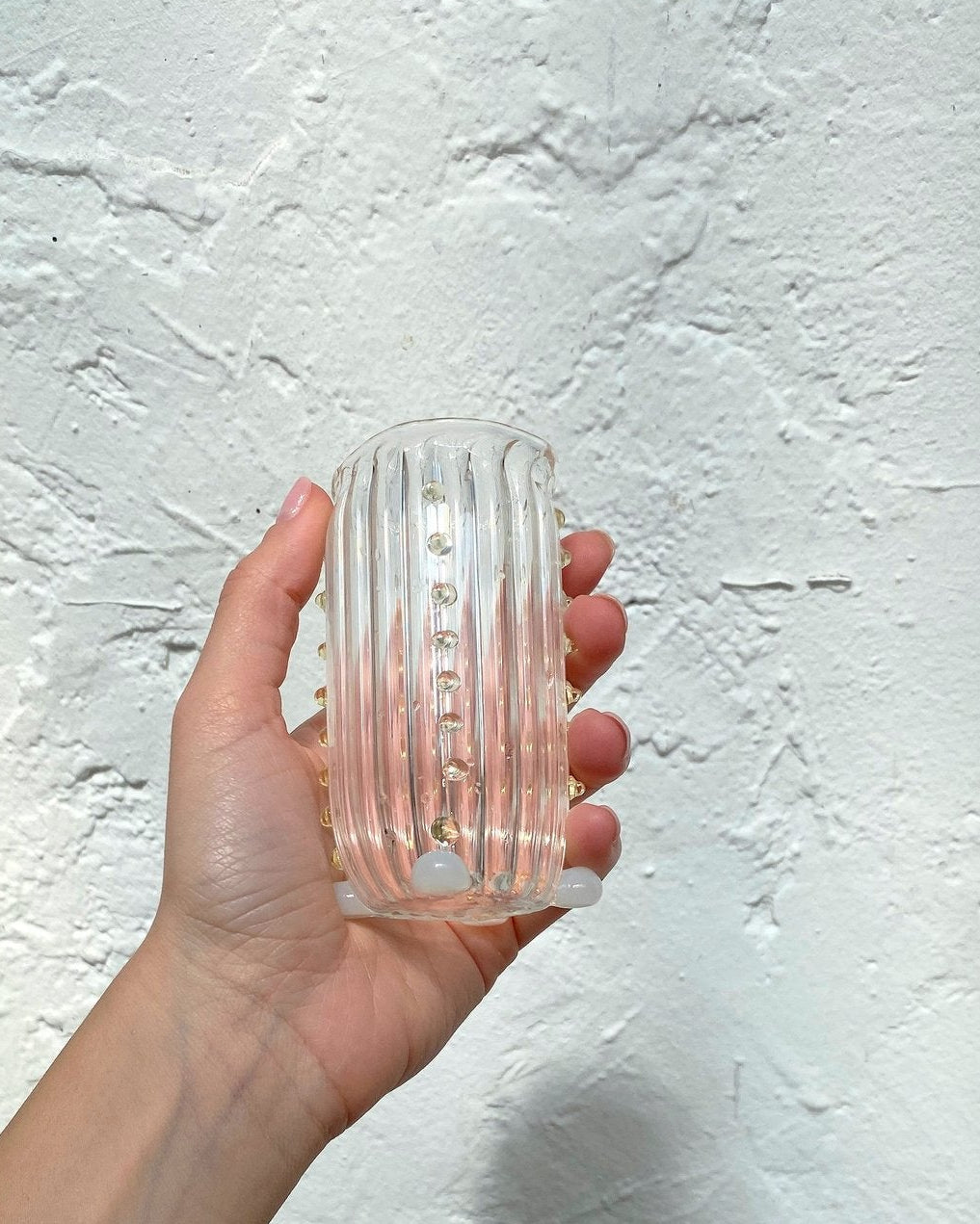 Somebody holding a mouth-blown, decorative water glass by Vivian Alarcón against a white brick wall wearing a yellow shirt.
