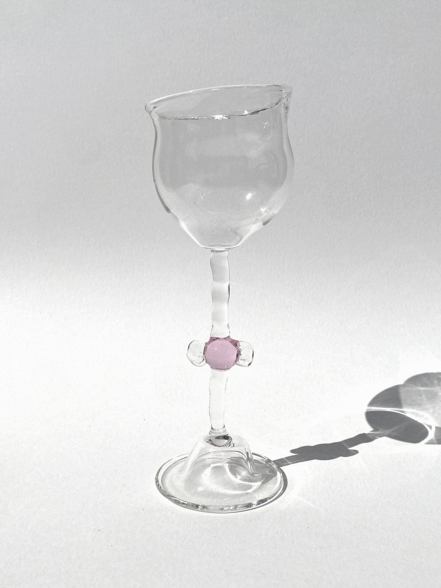A clear mouth-blown wine glasses by Vivian Alarcón with a pink candy-shaped embellishment on the stem, displayed against a light background.