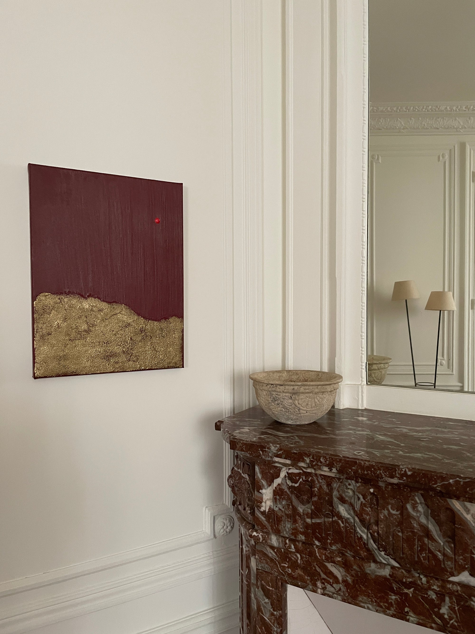 Red and gold abstract art by Noelia Hobeika on a white wall, besides a marble fire that has a stone decorative bowl positioned on top. There's a mirror above the fire place which shows a floor standing lamp in the reflection.
