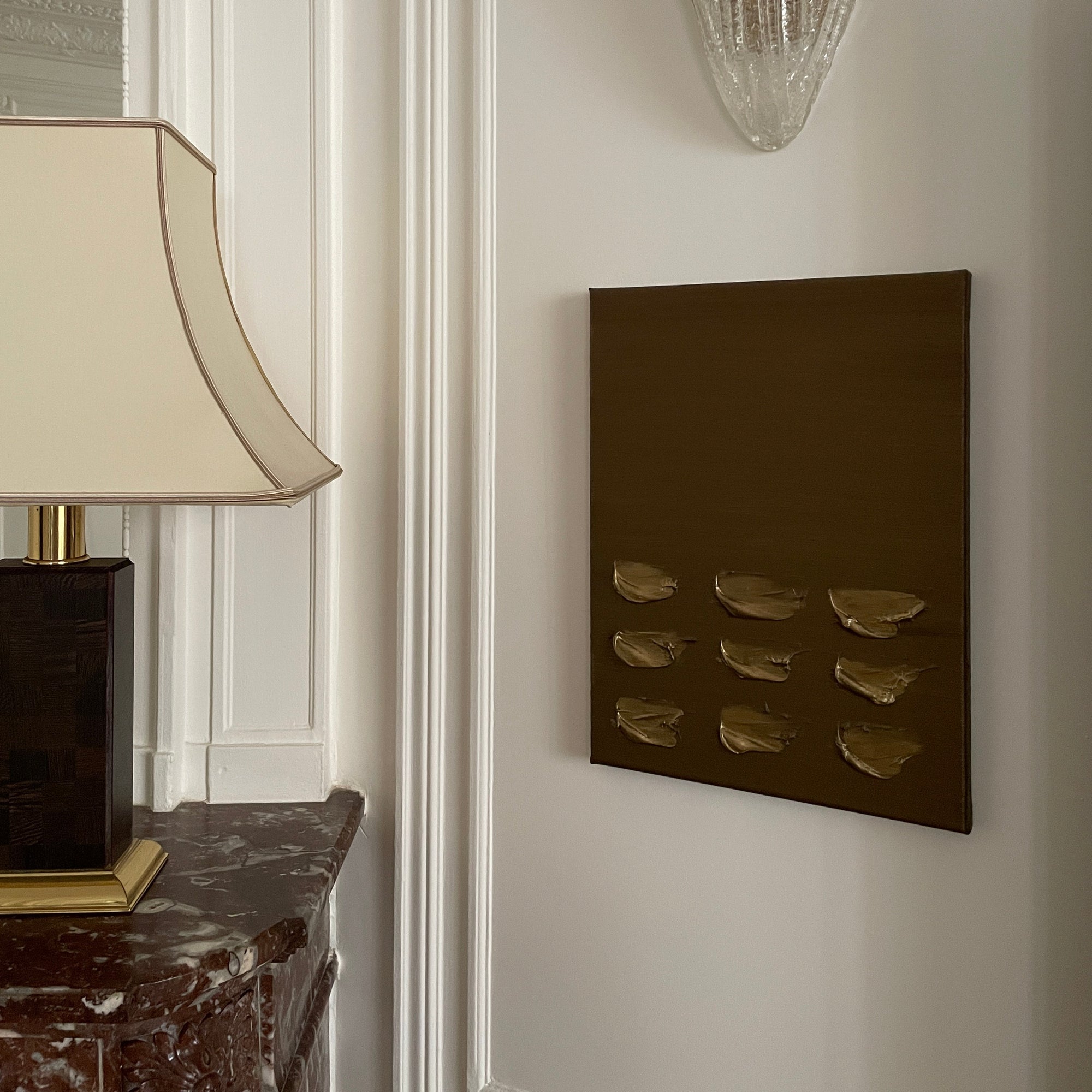 Gold and brown abstract wall art by Noelia Hobeika, on a white wall positioned below a decorative wall sconce and besides a lamp that is placed on top of a marble fire.