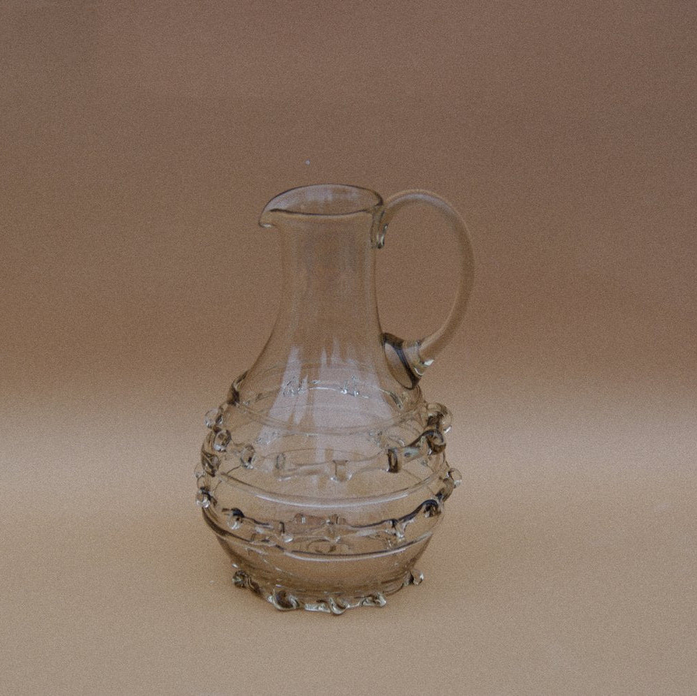 Smokey grey carafe with ribbed glass decorative accents on the base and a glass handle by Lucie Claudia, on a beige background.
