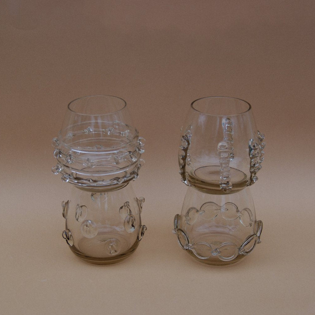 Four smokey grey water glasses with glass decorative accents by Lucie Claudia on a beige background.