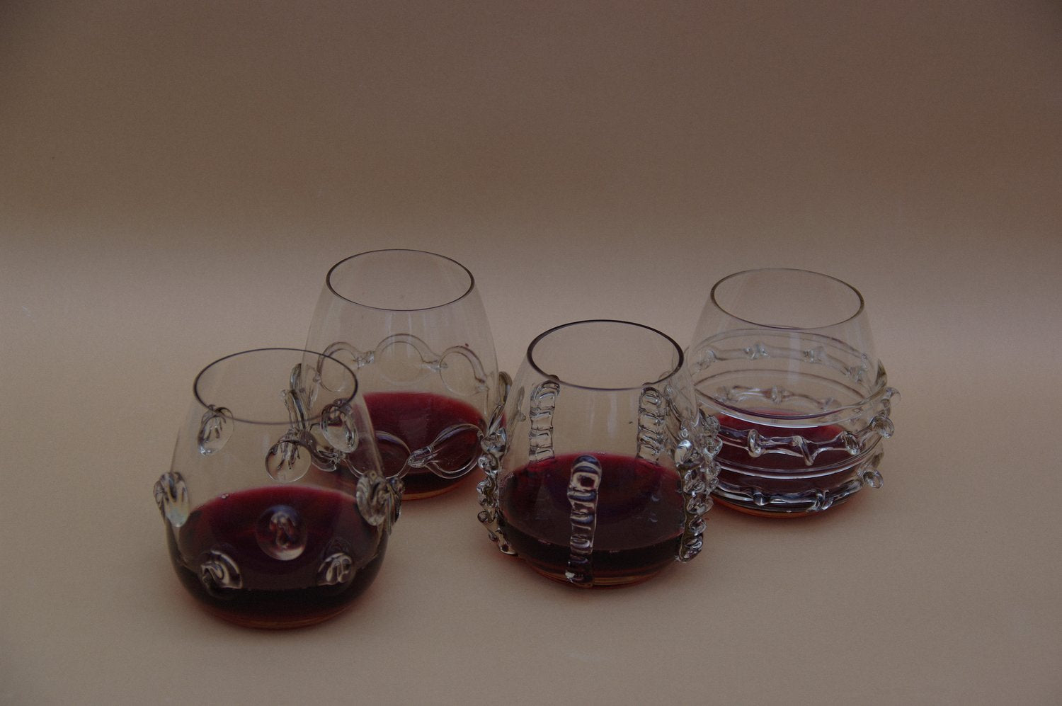 Four smokey grey water glasses with glass decorative accents by Lucie Claudia filled with red wine on a beige background.