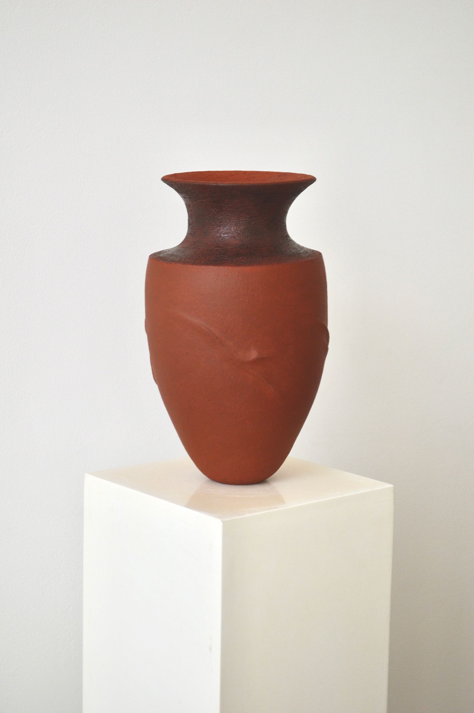 Terra cotta clay vase by Zeynep Boyan on a white pedestal against a light grey background.