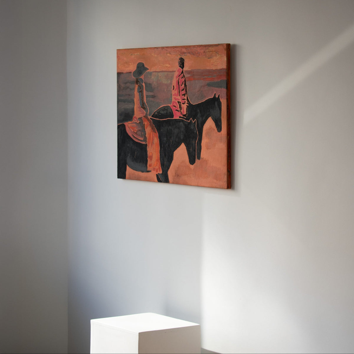 An original painting by Marcsen, depicting two riders on horseback in warm orange, red, black, and brown tones, displayed on a neutral wall above a white pedestal.