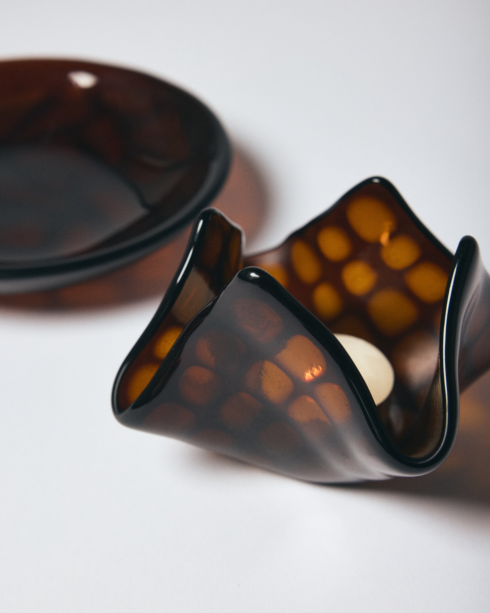 Close up of dark amber patterned glass candle holder with candle lit inside and matching glass bowl by Savoia Everything on a white background. 
