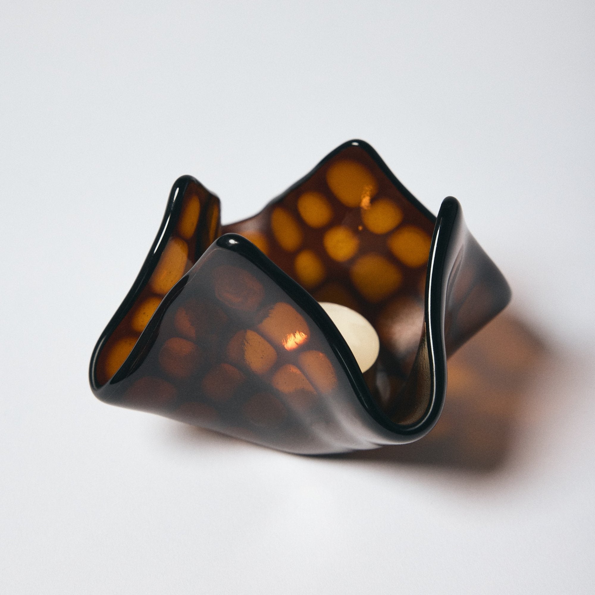 Dark amber patterned glass candle holder by Savoia Everything with candle lit inside on a white background.