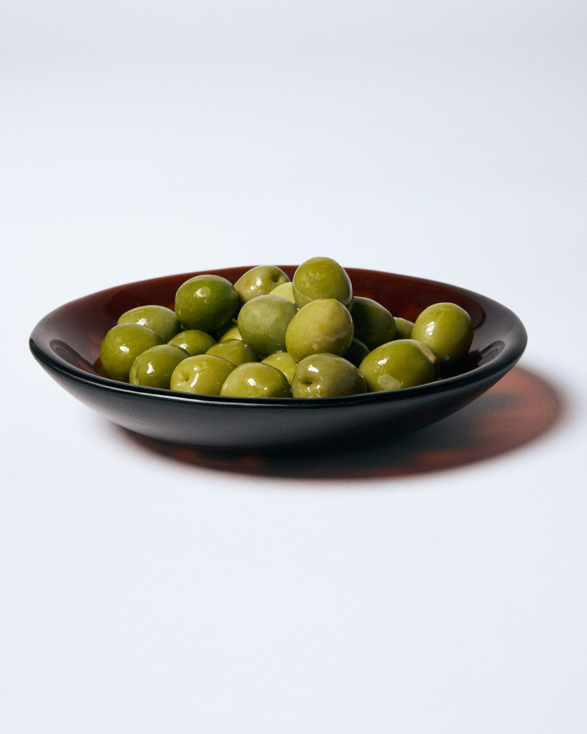 Dark amber glass bowl holding green olives by Savoia Everything on a white background.