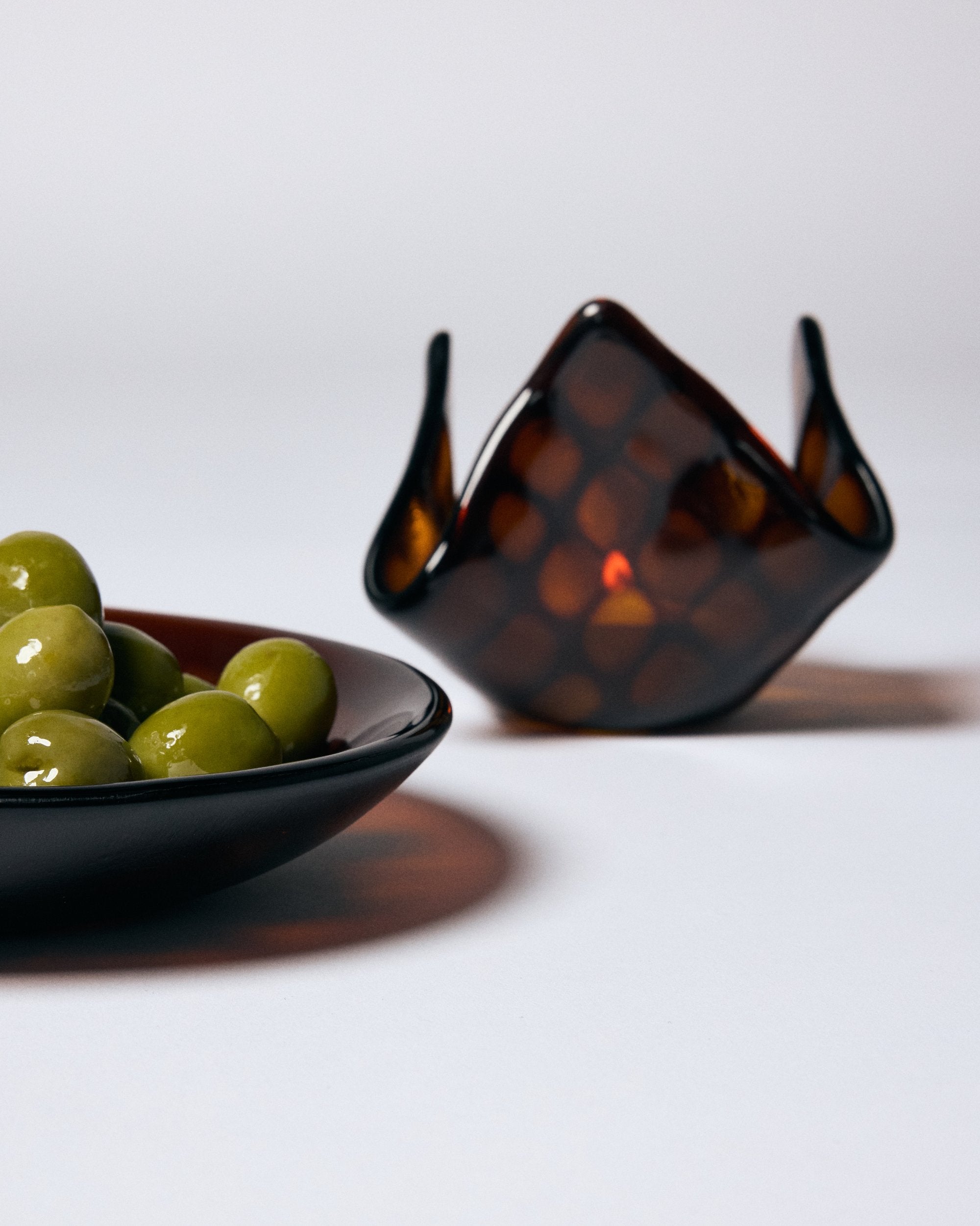 Dark amber glass bowl holding green olives. In the background is the matching candle holder with a lit candle by Savoia Everything on a white background.