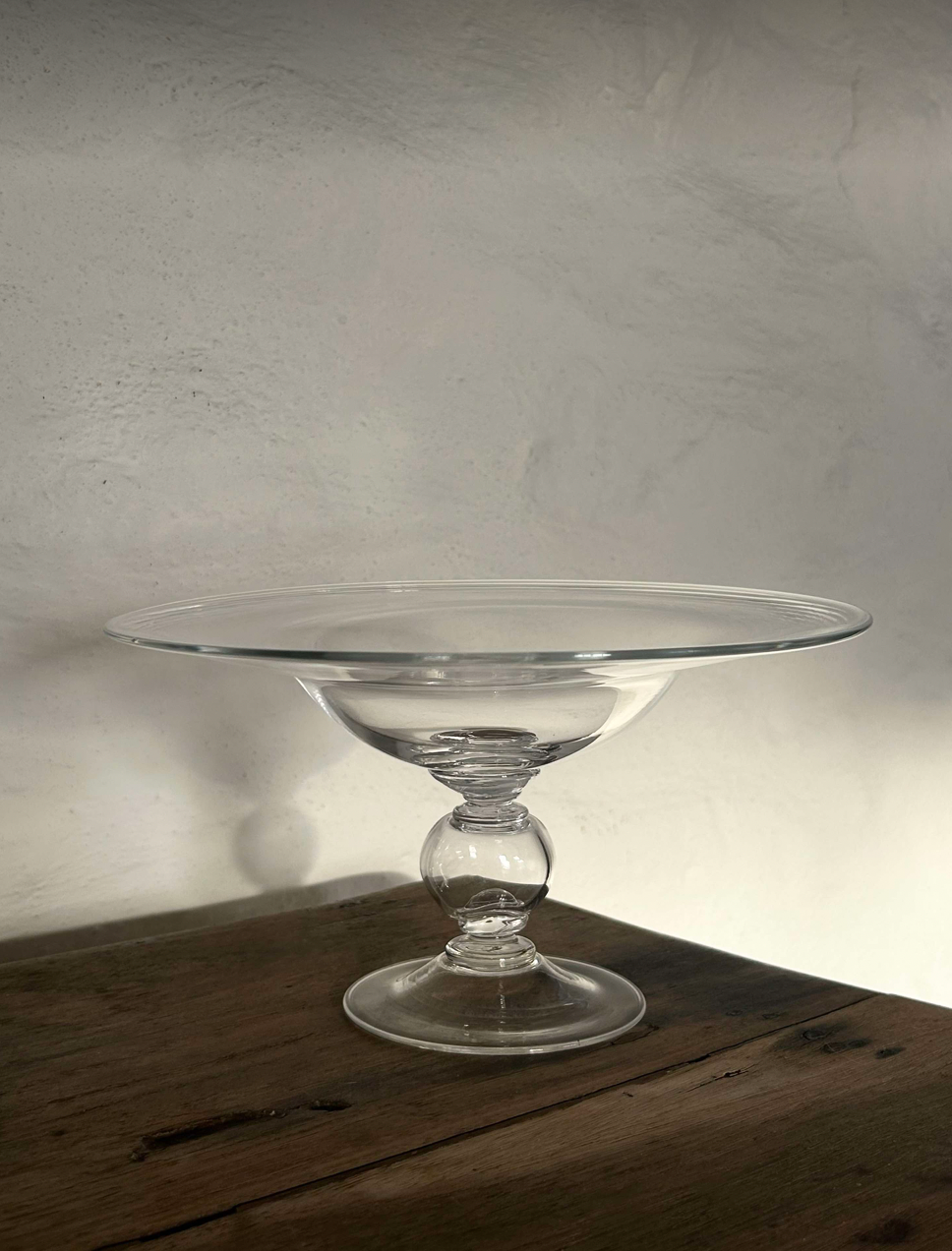 Clear glass footed bowl by Bastian Bjerge on a wooden table with a neutral painted wall. 