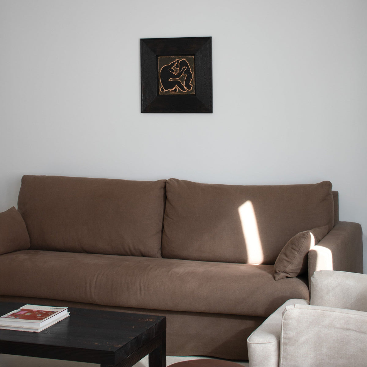 A brown fabric sofa in a minimalist living room setting, with framed original wall art by Marcsen hanging on the white wall above it.