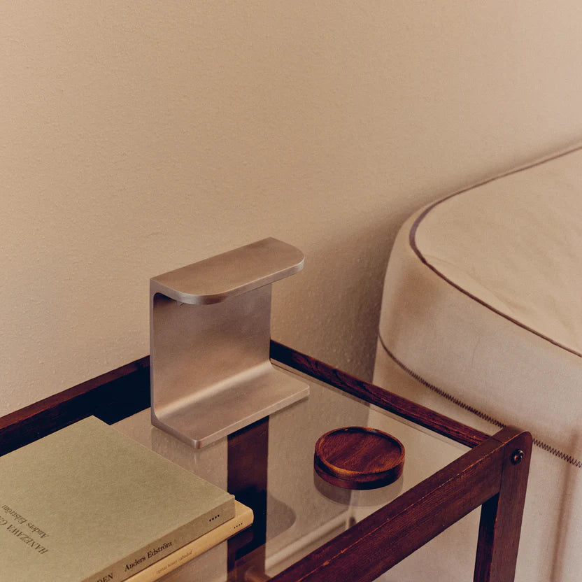 Desk lamp by PARCTA with the light off on a glass bedside table that has wooden accents, besides a bed with cream linen. On the beside table is also two books and a wooden coaster.