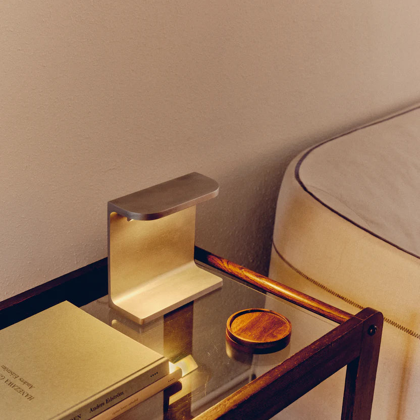 Desk lamp by PARCTA with the light on on a glass bedside table that has wooden accents, besides a bed with cream linen. On the beside table is also two books and a wooden coaster.