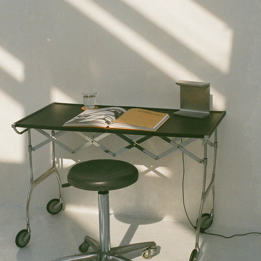 Desk lamp by PARCTA with the light off placed on an industrial desk with a leather stool. On the desk is an open magazine and a glass of water.