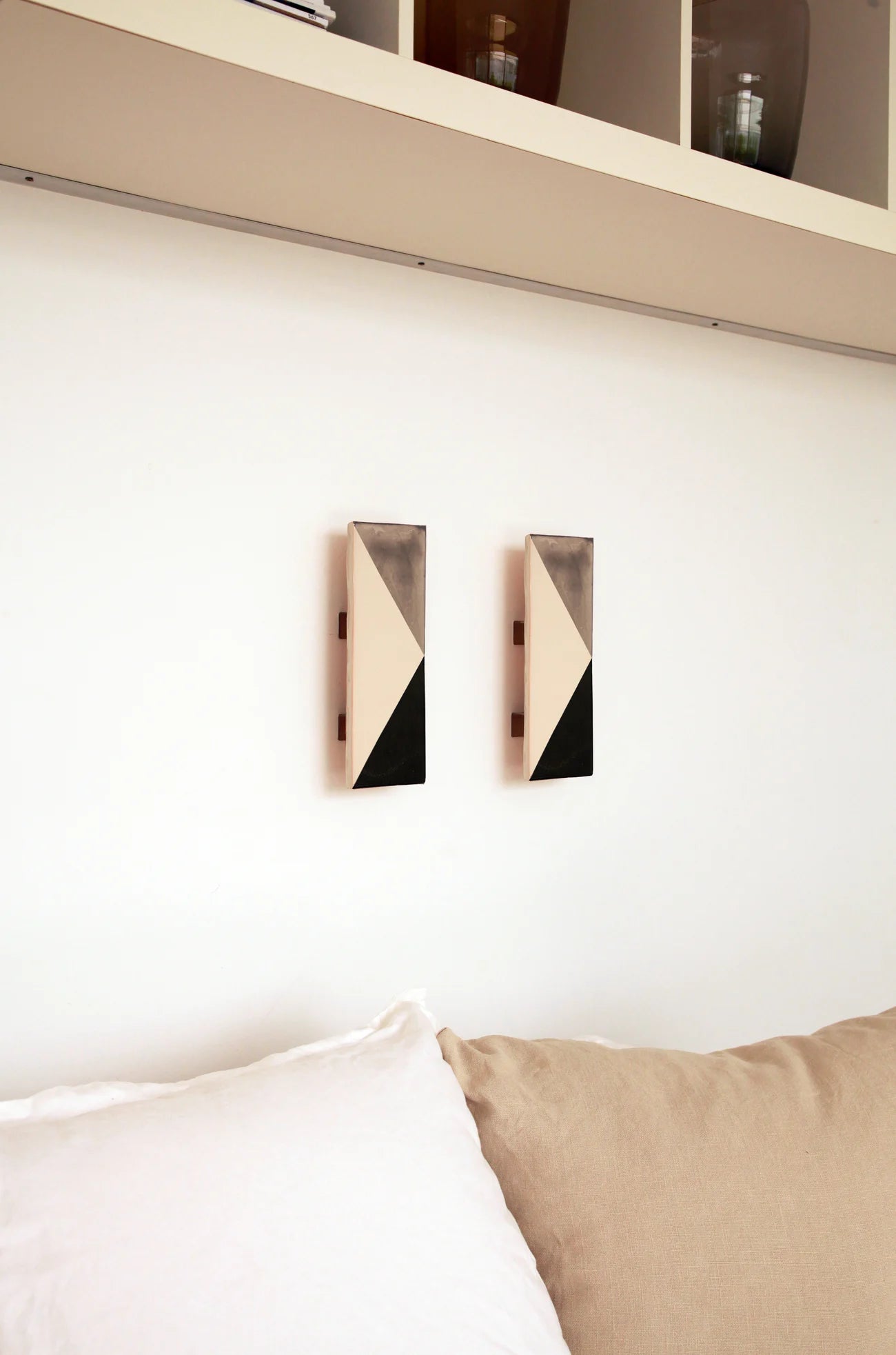 Two wall-mounted light fixtures by Violaine d'Harcourt with the light off, placed below a shelf and above a sofa. The design is a geometric pattern in grey, black and white.