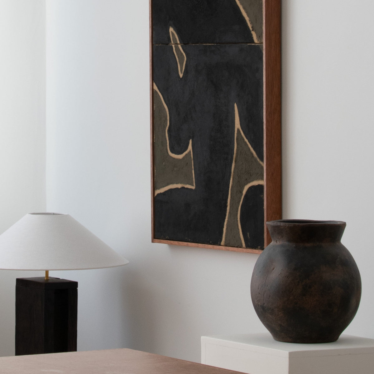 Original abstract wall art by Marcsen in black and earthy green tones housed in a vertical wooden frame, next to a black table lamp and a dark ceramic vase on a white pedestal.